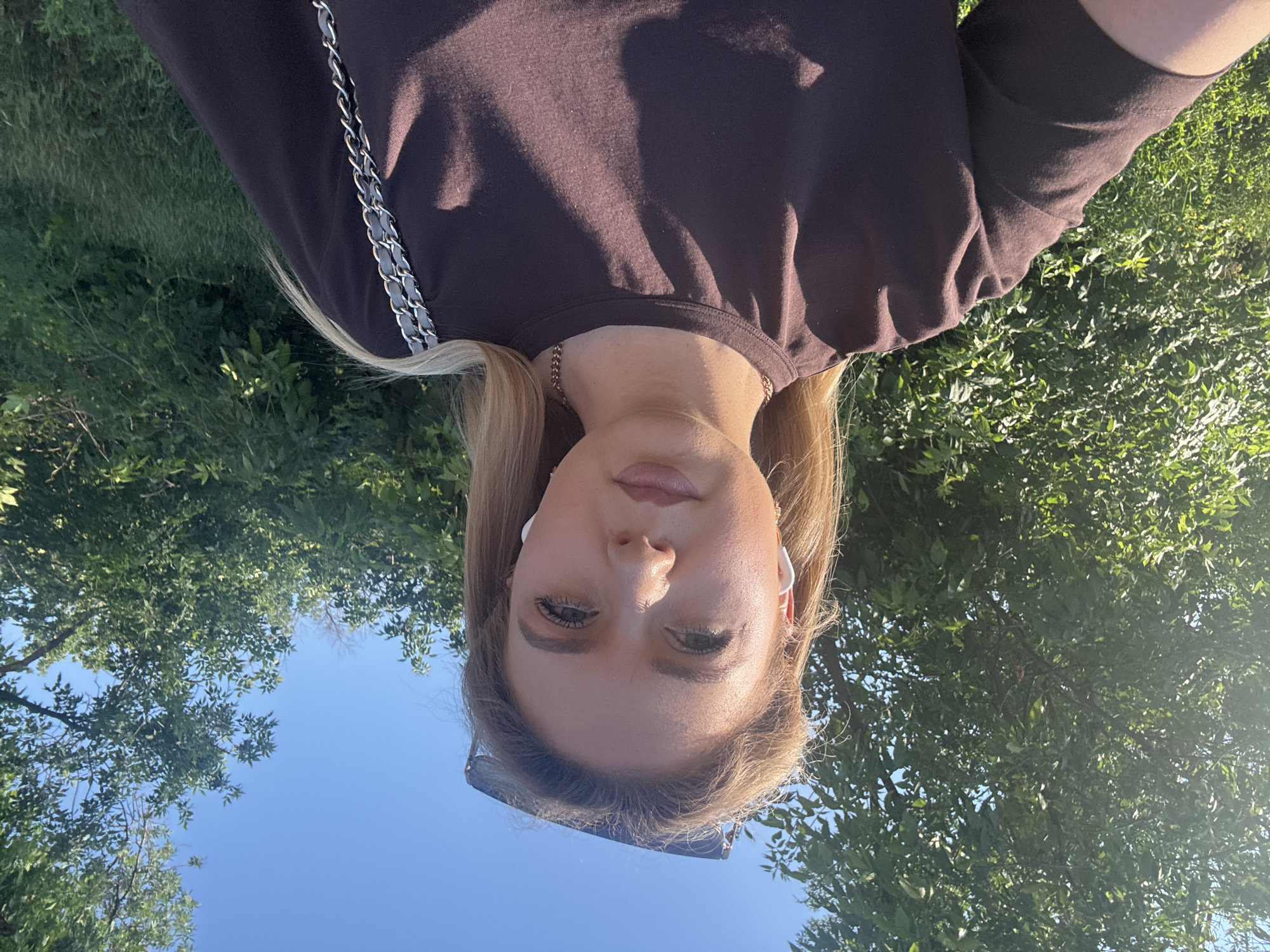 A young woman with blonde hair and white earbuds takes a selfie outdoors. She is wearing a dark brown t-shirt and a silver chain strap bag slung over her left shoulder. Sunglasses are perched on top of her head. She is standing in front of lush green trees and grass, with a clear blue sky above. The scene appears to be set in a park or a natural area, possibly in the city of Unknown, Unknown, during daylight hours, as indicated by the bright sunshine.