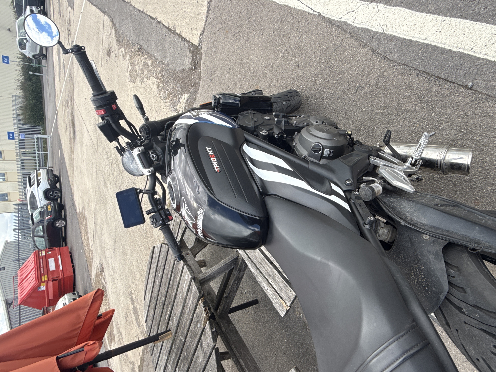 The image shows a black Triumph Trident motorcycle parked next to a wooden picnic table, with an orange umbrella overhead. In the background, there is a red skip bin, a dark-colored car, and a white SUV. Further back, there are industrial-looking buildings and a fence. The scene appears to be an outdoor parking lot or commercial area, possibly near a business or industrial estate in Tewkesbury, United Kingdom. There are no people visible in the image. The motorcycle is the primary subject, positioned at an angle to showcase its features. The environment is overcast and appears to be a paved area with some rougher surfaces and white line markings.