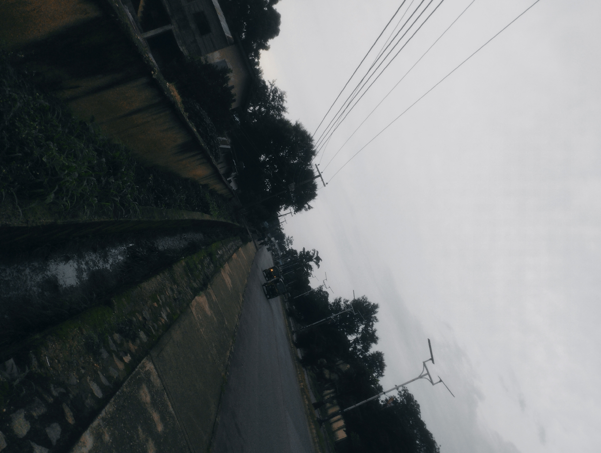 The image, captured from an elevated perspective, shows a street scene in Jos, Nigeria. The orientation is tilted, with the sky occupying the upper right quadrant and a roadside drain occupying the lower left. The drain is partially filled with water and lined with concrete and stones. Adjacent to the drain is a paved walkway or sidewalk. The street itself is asphalt, with a light sheen suggesting it might be wet. Two three-wheeled vehicles, commonly known as Keke Napeps, are visible on the street, one with its headlights on. Trees with dense foliage line the right side of the street, and electrical wires are strung overhead, supported by utility poles. In the background, on the left side, are the roofs and upper portions of buildings, some appearing weathered and partially obscured by vegetation. The overall atmosphere is overcast, indicated by the uniformly gray sky.