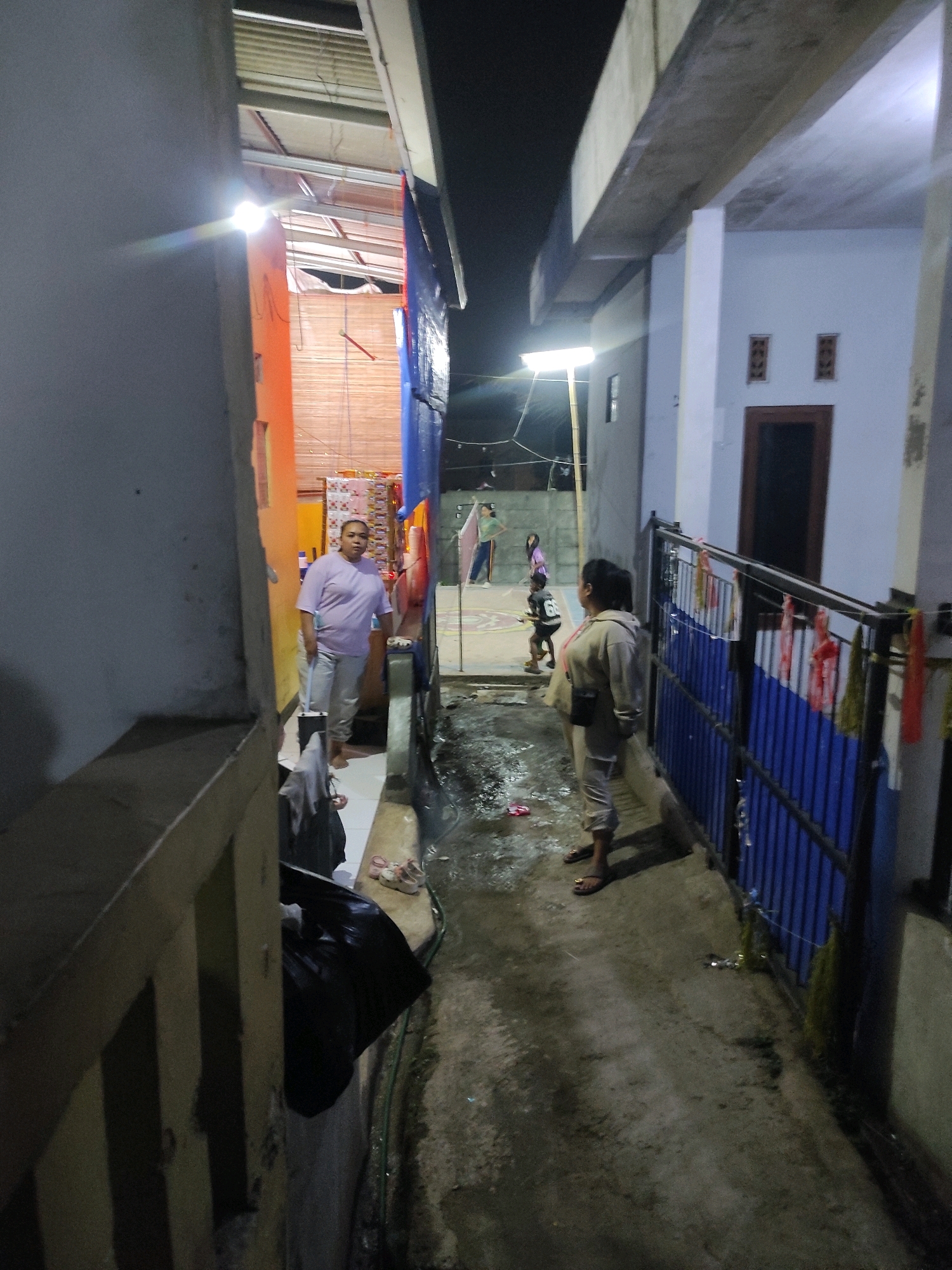 The image depicts an alleyway in what appears to be a residential area of Padalarang, Indonesia, at night. A woman wearing a light purple t-shirt and light-colored pants is standing in the alley, holding a blue mop handle. To her left, a large black garbage bag is placed against a wall. Further down the alley, a person wearing a light-colored jacket and pants stands facing away from the camera, near a blue fence with red and white fabric draped over it. In the background, several other individuals, including children, are visible on what looks like a basketball court or a paved recreational area. The ground of the alley is wet and reflective, suggesting recent rain or cleaning. Lighting in the scene comes from overhead lamps and ambient sources, casting shadows. The architecture suggests simple, possibly unfinished, structures.