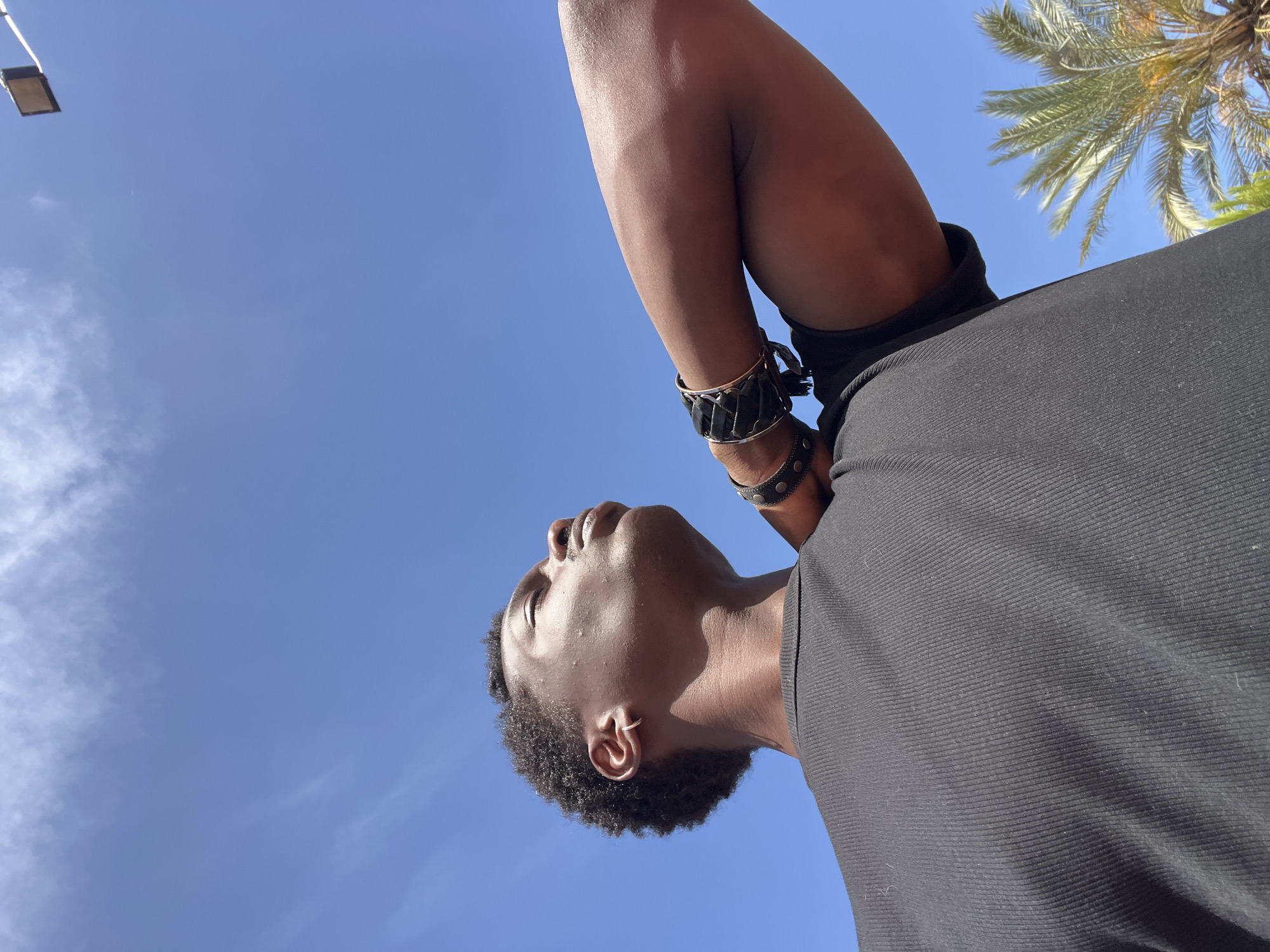 A low-angle shot shows a Black man from the chest up, looking up towards a clear blue sky. He wears a black ribbed t-shirt and has his right arm bent and resting behind his head. He has dark, curly hair and a thin hoop earring in his left ear. He is wearing several bracelets on his right wrist, which appear to be made of leather and metal. Sunlight highlights his skin and the contours of his face and arm. In the upper right corner of the image, a light fixture with a square brown shade is visible, attached to a white pole against the sky. In the lower right, the fronds of a palm tree are visible. The overall scene suggests an outdoor setting, possibly in a warm climate. The presence of the palm tree and the light fixture is consistent with an urban outdoor public space, like a park or plaza, potentially in a Mediterranean city. Given the location of Sant Adrià de Besòs, Spain, it is plausible that this image was captured in one of its public recreational areas or along its coastline, where such lighting and vegetation are common.