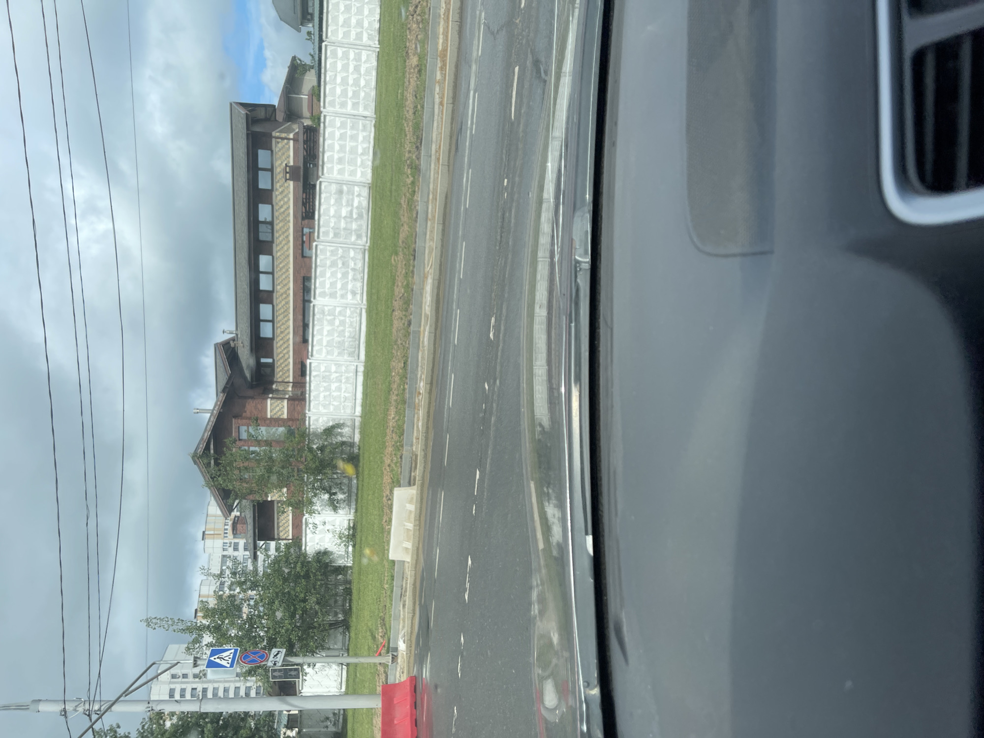 The view is from inside a car, looking out the passenger-side window at a street scene. On the left, there is a multi-story brick building with numerous windows, adjacent to a section of what appears to be decorative concrete block wall. Beyond the building, there are power lines and a pedestrian crossing sign. The road next to the car shows white lane markings, and there is grass alongside the road. The presence of a Russian pedestrian crossing sign suggests the location is in Moscow, Russia.