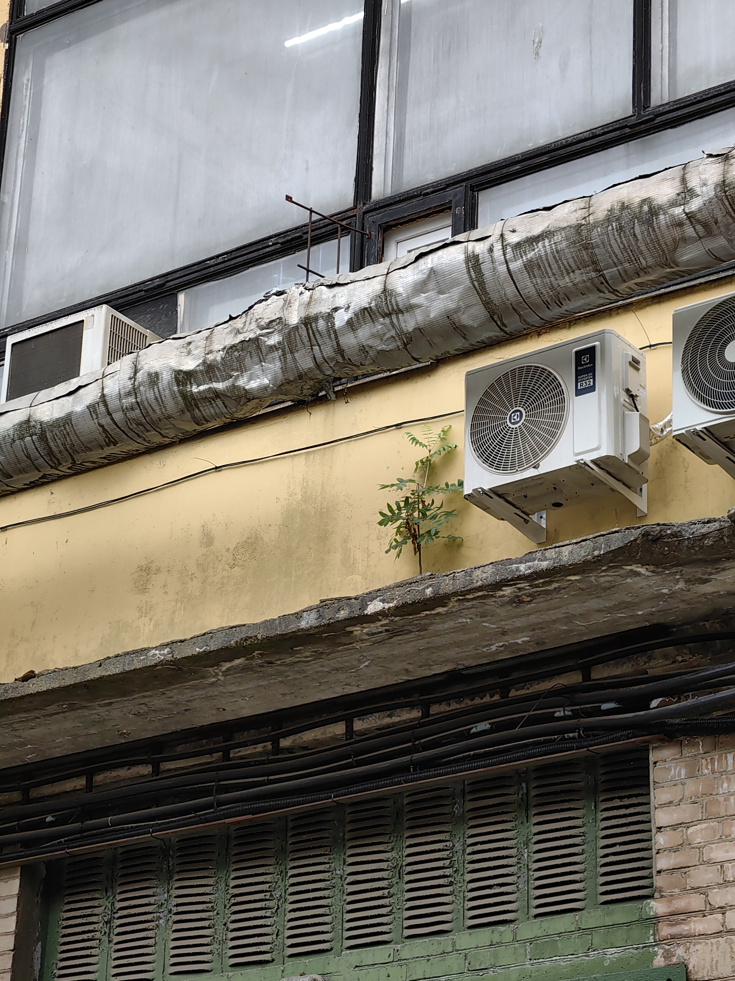 The image shows the exterior of a building with yellow-painted walls. Two white air conditioning units are mounted on the wall, one with the brand "Electrolux" visible. A small tree with green leaves is growing from a ledge below the air conditioning units. Above the yellow wall, there is a large, possibly frosted window and a large insulated pipe with dark streaks running along it. Below the ledge, there are several black pipes and a green louvered vent on a brick wall. The scene depicts an urban environment, possibly in Minsk, Belarus, characterized by utilitarian building design and the presence of building infrastructure.