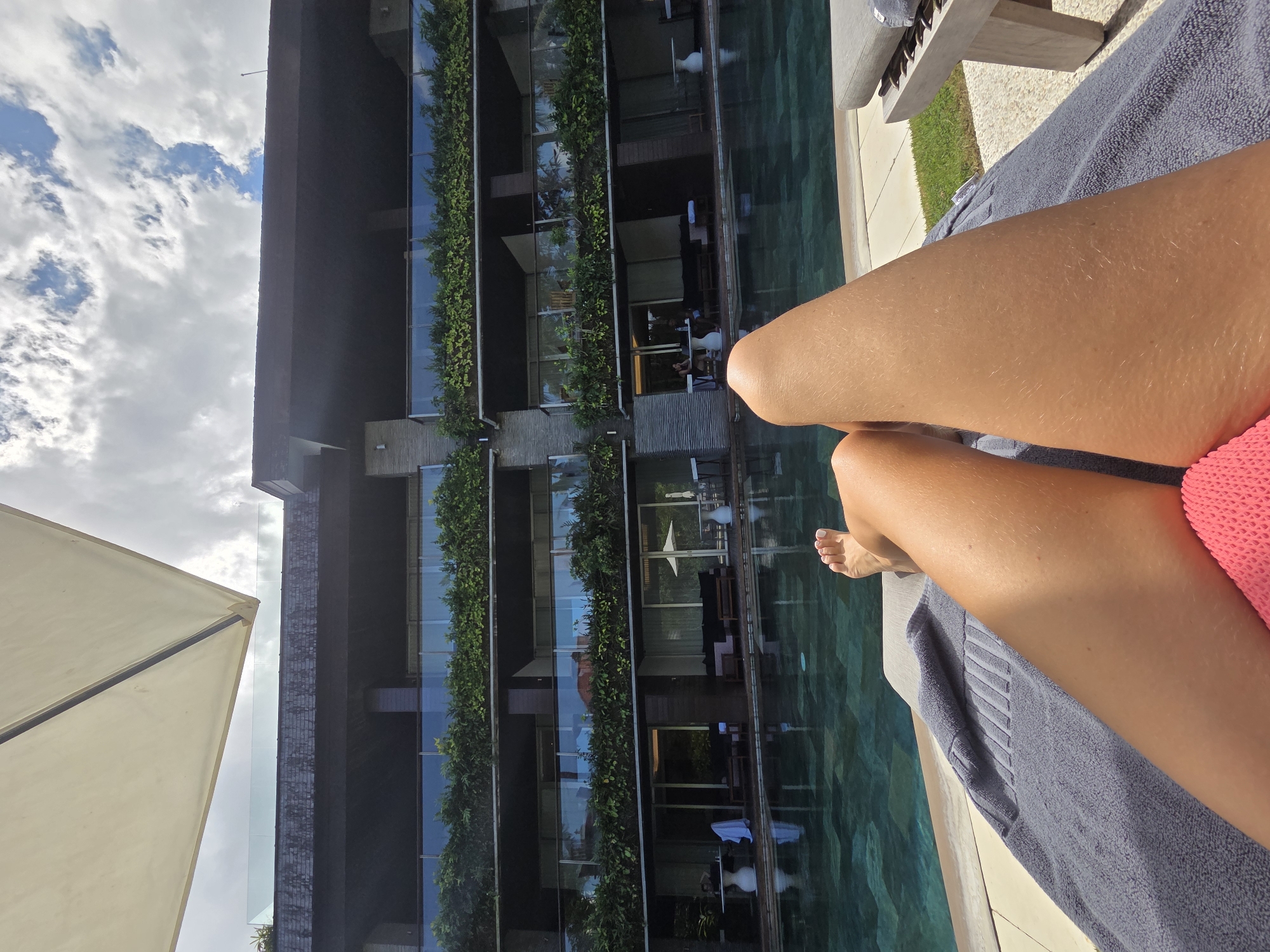 The image depicts a woman sunbathing next to a pool at the COMO Uma Canggu hotel in North Kuta, Indonesia. The perspective is from above, focusing on the woman's legs in the foreground, partially covered by a gray towel on a lounge chair. She is wearing a pink bathing suit. The pool is adjacent to the chair, reflecting the building across from it. The building has multiple levels with balconies adorned with greenery. In the background, the sky with scattered clouds is visible. Part of a white parasol is visible in the lower left corner.