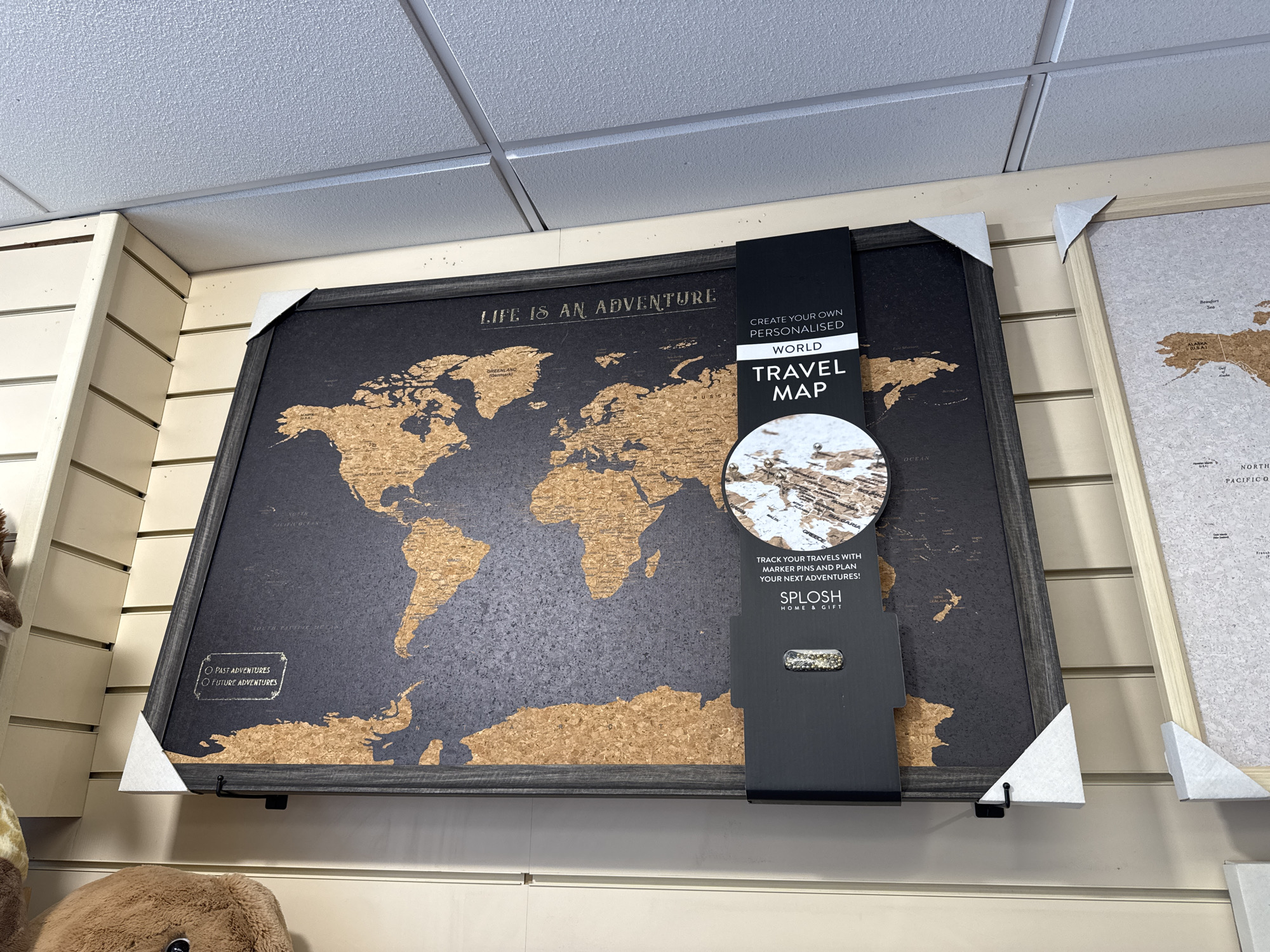 The image shows a close-up of a personalized world travel map made of corkboard, displayed on a slatted wall in a retail environment. The map is framed in dark wood and features the text "LIFE IS AN ADVENTURE" at the top. A black card attached to the right side of the map reads "CREATE YOUR OWN PERSONALISED WORLD TRAVEL MAP" and mentions "TRACK YOUR TRAVELS WITH MARKER PINS AND PLAN YOUR NEXT ADVENTURES!" Below this, the brand "SPLOSH HOME & GIFT" is visible, along with a small clear plastic container holding gold-colored pins. The map itself shows continents in cork material against a dark grey background, with some country names faintly visible. To the right of this map, another similar corkboard world map is partially visible, framed in light wood. The background consists of the slatted wall and a suspended ceiling. The scene is well-lit, suggesting an indoor retail space. No specific location in Clonmel, Ireland is indicated by any visible text or landmarks.
