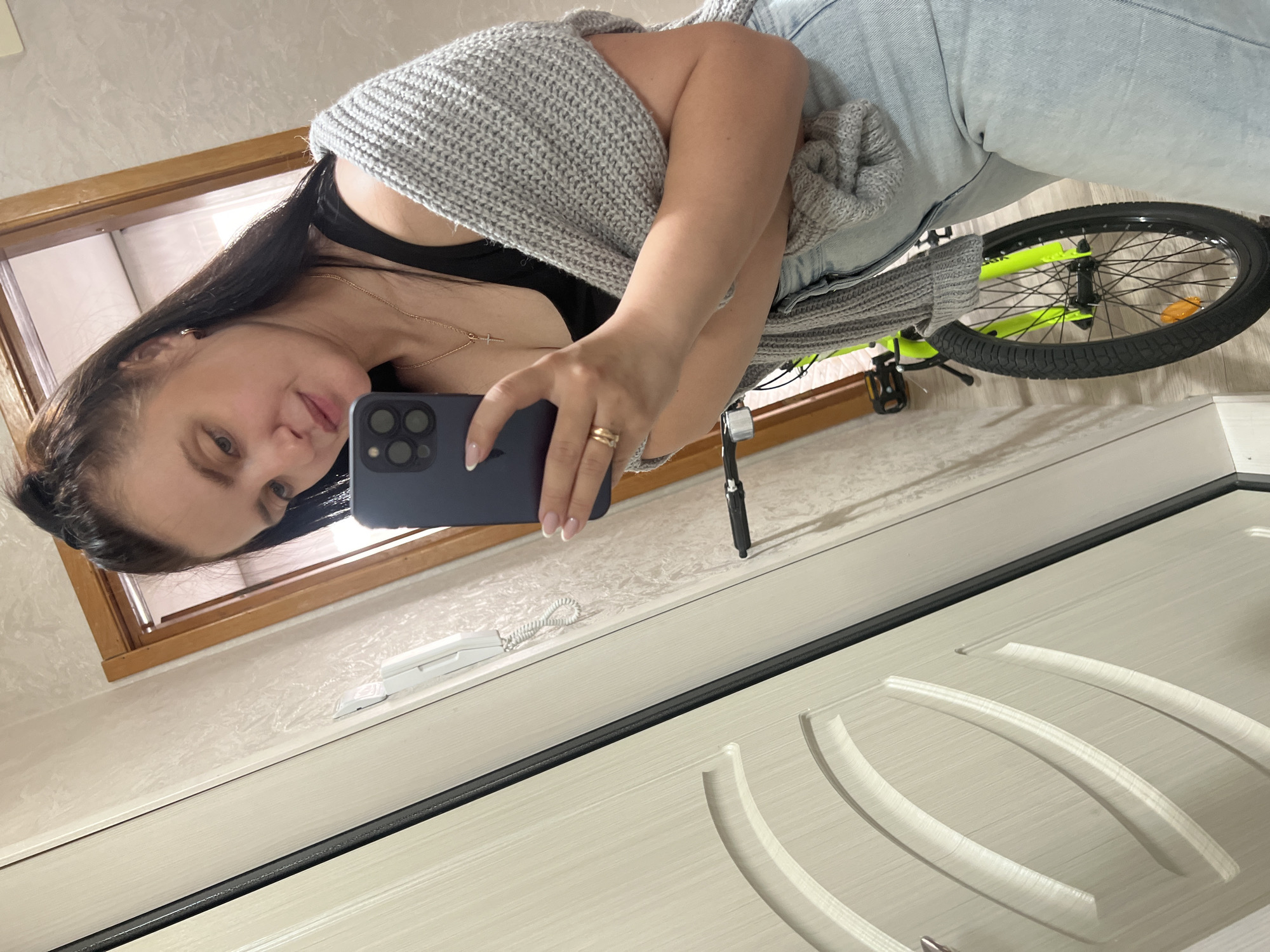 The image is oriented sideways, depicting a full-length mirror selfie of a woman taken indoors. The woman, with long dark hair, is positioned vertically in the center-left of the frame, facing the camera's reflection. She is wearing a black top, a textured grey knitted cardigan, and light blue jeans. She holds a dark-colored smartphone, likely an iPhone given its camera array, in her right hand to capture the image. A gold-colored ring is visible on her right ring finger, and her fingernails feature white French tips. Her expression is calm with a slight smile.
To the right of the woman, a bright fluorescent green/yellow bicycle is partially visible. Its front wheel, with black tires and spokes, and part of its frame are seen. A black pedal is also identifiable. The bicycle rests on a light-colored wooden floor.
The background includes a wall with light-colored, subtle textured wallpaper. A brown wooden frame, possibly from a window or an internal doorway, is visible in the upper left corner of the image. A white intercom or doorbell unit with a coiled cord is mounted on the wall. A prominent light-colored door with decorative, carved curved panels and a black horizontal trim line occupies the lower and right parts of the frame.
The image is a reflection, giving a reversed perspective of the scene. There is no clearly legible text visible in the image.