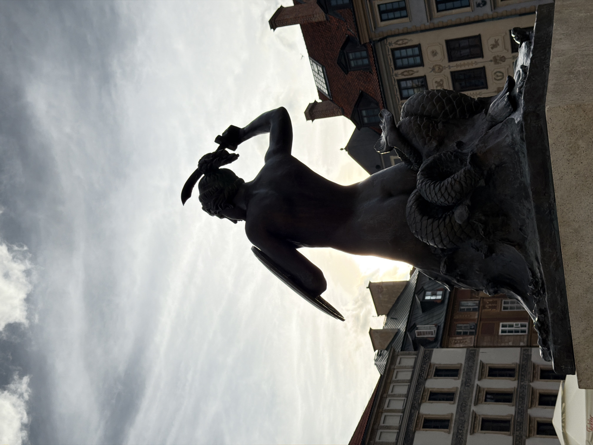 The image shows the Mermaid of Warsaw statue, a bronze sculpture located in the Old Town Market Square (Rynek Starego Miasta) of Warsaw, Poland. The statue depicts a mermaid holding a sword and a shield, poised as if in defense. The sculpture is dark bronze and is set on a stone pedestal. In the background, traditional Warsaw architecture with red tiled roofs and cream-colored facades is visible. The sky above is cloudy and overcast, with the sun attempting to break through, creating a soft illumination. The angle of the photograph is from below the statue, looking upwards.