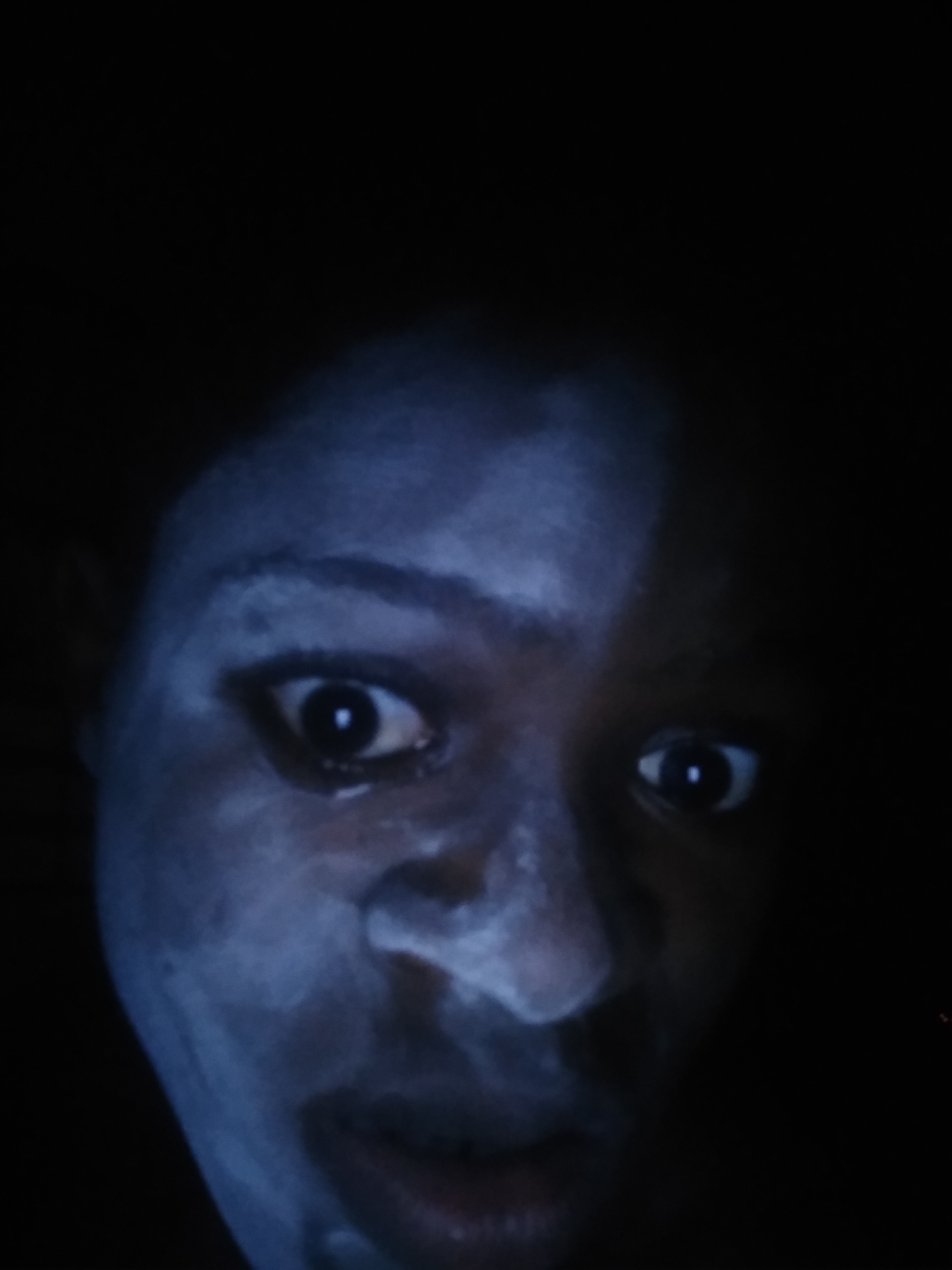 A close-up image of a person's face, heavily shadowed with what appears to be a white or light-colored substance on the skin, potentially a mask or covering. The person's eyes are wide and visible. The image is low-lit. There is no indication in the picture about the location in Shangisha, Nigeria.