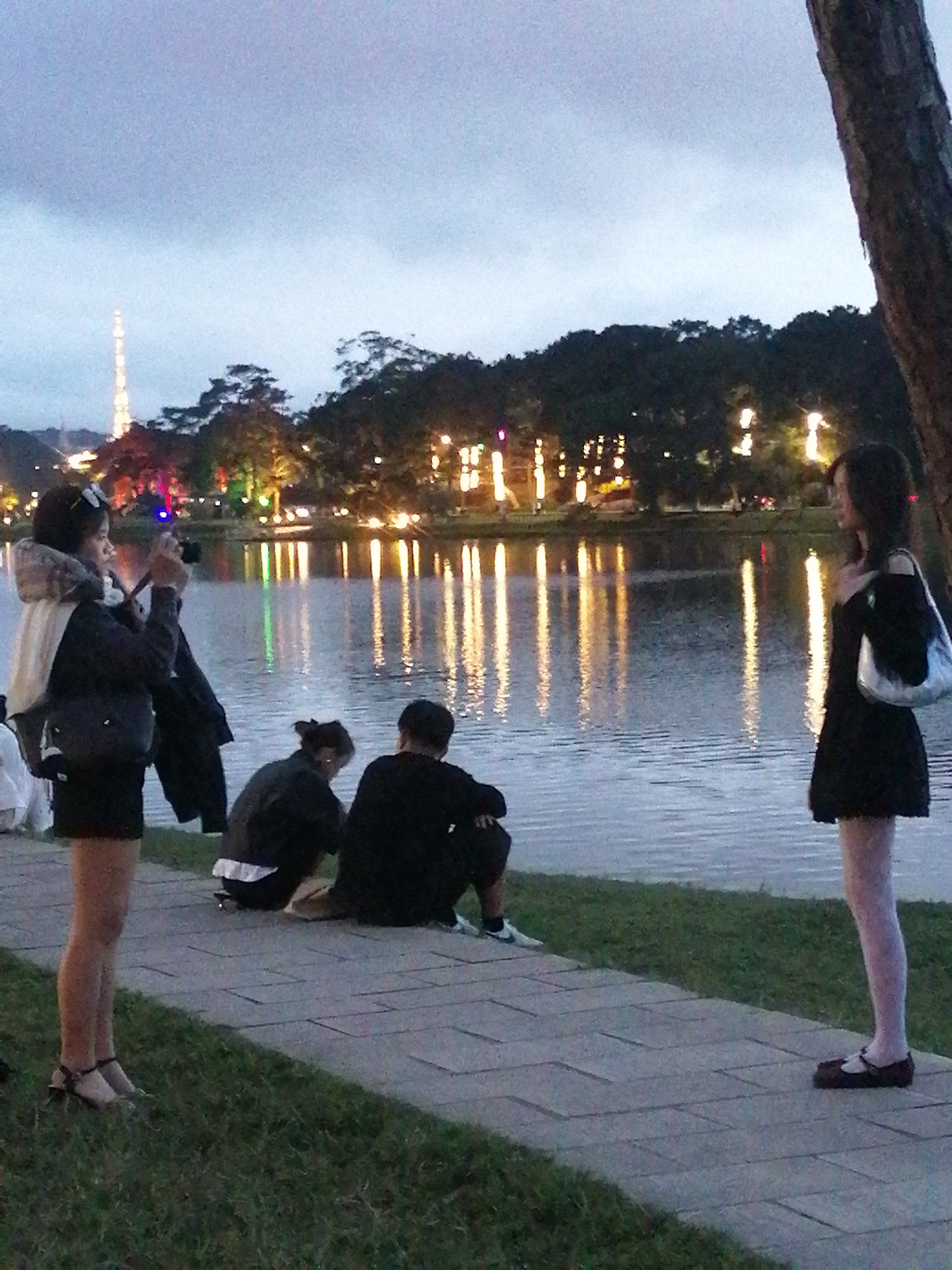 In Da Lat, Vietnam, at dusk, four individuals are gathered near the edge of a tranquil lake. A woman in the foreground on the left, wearing a black jacket with a fur-lined hood and a black skirt, is actively taking a photo with a camera. To her right, two people are seated on the ground, facing away from the camera, their backs to the lake. Further to the right, a woman in a black dress and white tights stands with her back to the lake, looking towards the left, and holding a light-colored handbag. The background features a tree line with illuminated buildings and lights reflecting on the water, suggesting an urban park or recreational area by the lake. The sky is overcast, contributing to the dim lighting conditions.