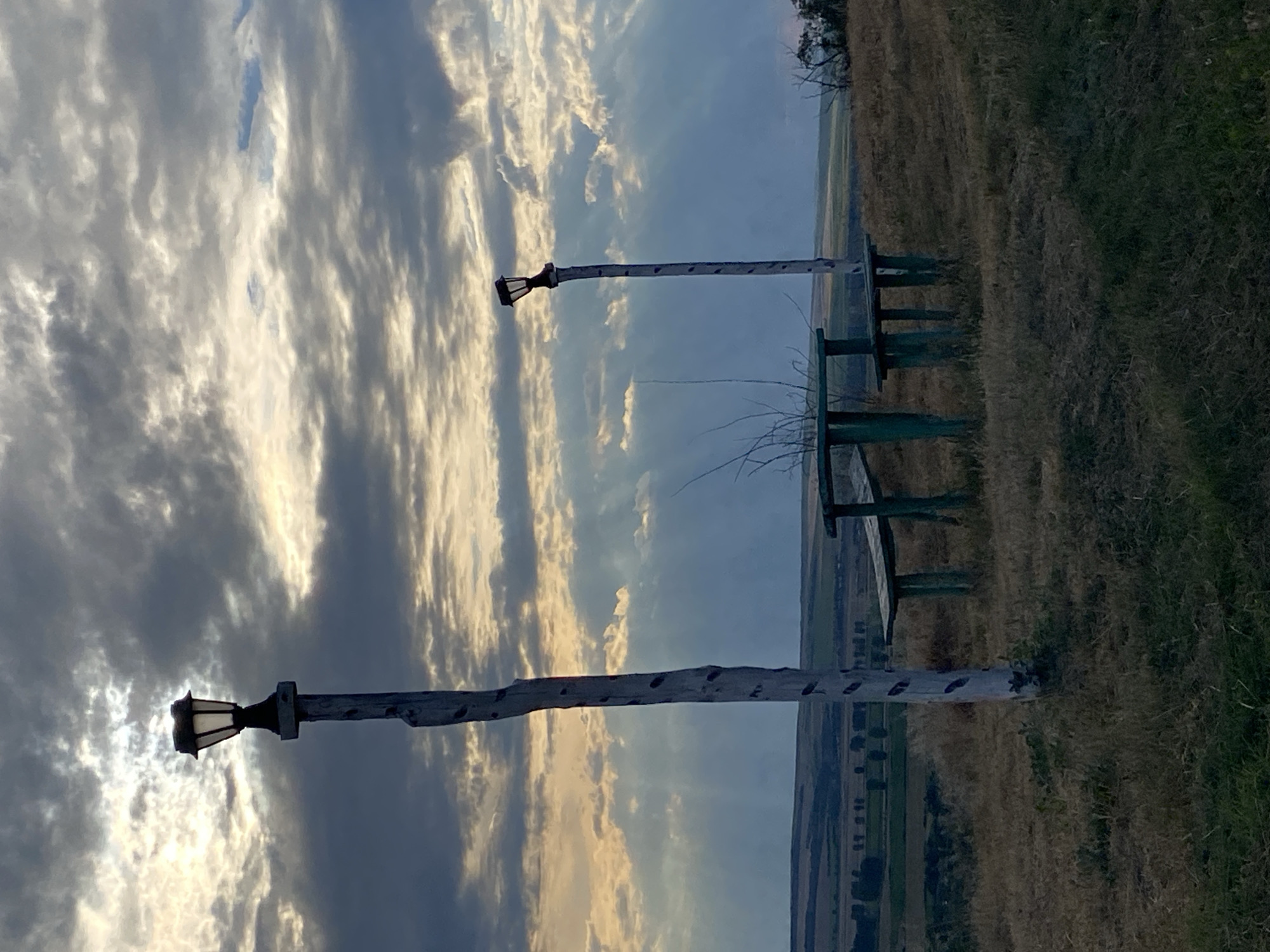 The image depicts an outdoor scene on a hill overlooking a rural landscape in Horești, Moldova. In the foreground, a rustic wooden picnic table with benches is positioned on dry grass and vegetation. Two tall, weathered wooden posts, each topped with a lantern, stand near the table. The sky above is dramatic with a mix of dark, heavy clouds and rays of sunlight breaking through, suggesting either dawn or dusk. The background shows rolling hills with fields and scattered trees. There are no people or animals visible in the scene.