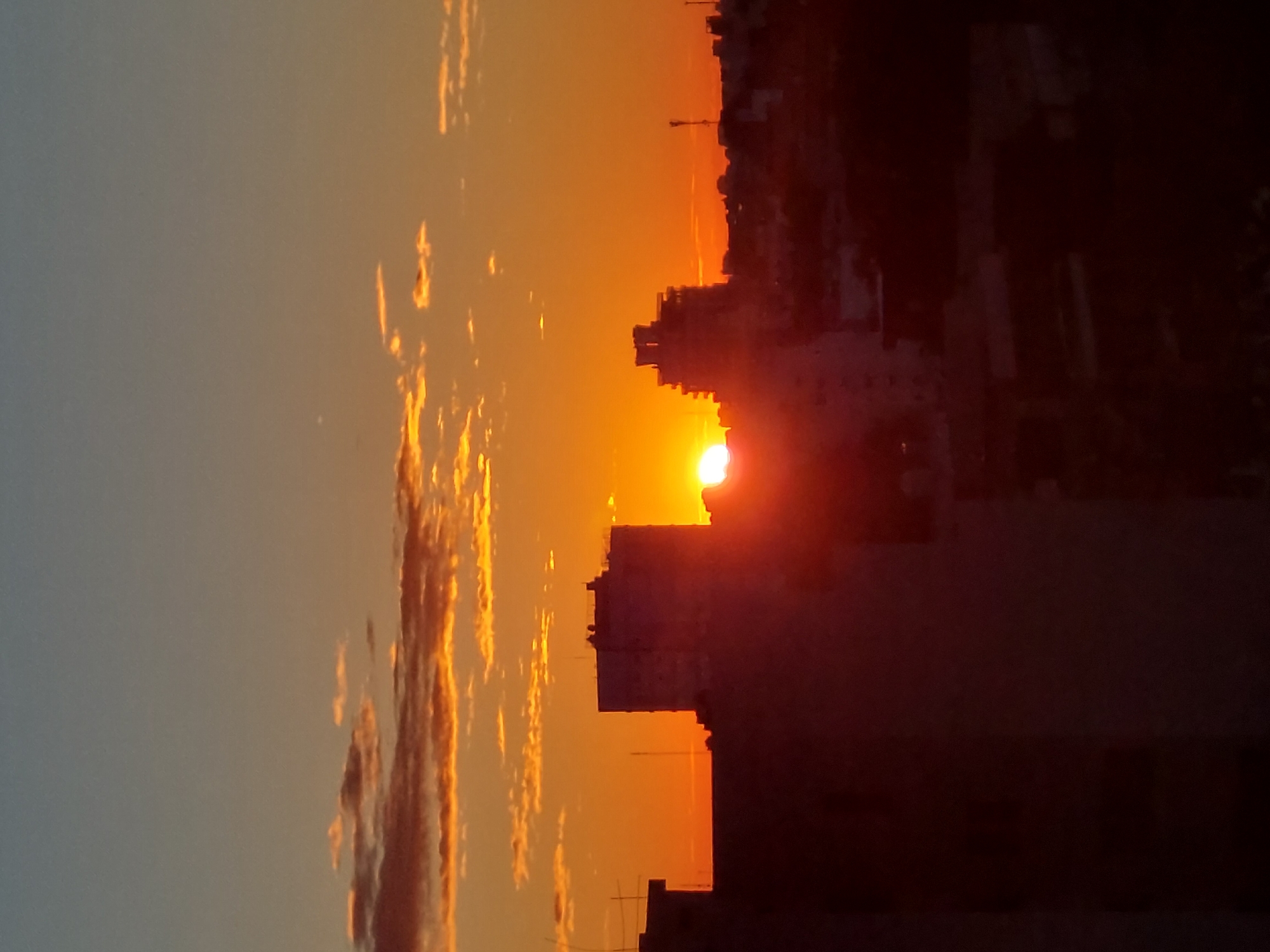 The image depicts a sunset over the skyline of Buenos Aires, Argentina. The sun is positioned low on the horizon, appearing between the silhouettes of buildings. The sky is filled with orange and golden hues, with the sun itself radiating a bright white light. In the foreground, the outlines of buildings are visible, creating a stark contrast against the vibrant sky. The scene suggests a clear day, with the warm light indicating the final moments of the day.