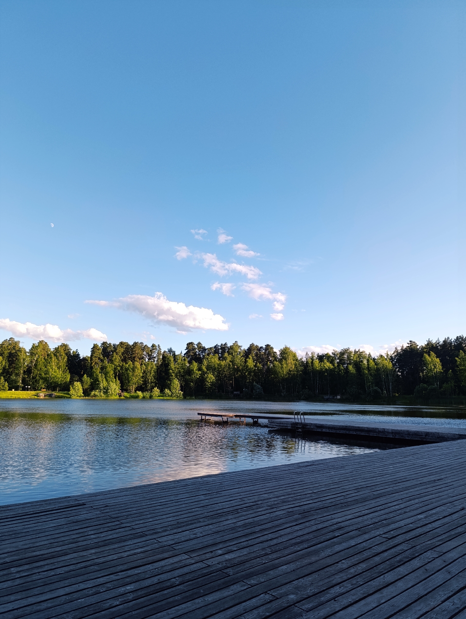 The image depicts a serene lake scene in Chernogolovka, Russia, under a clear, pale blue sky with a few wispy clouds and a visible crescent moon in the upper left quadrant. The time of day appears to be late afternoon or early evening, as indicated by the soft, diffused light.
In the foreground, a weathered wooden boardwalk stretches diagonally across the frame, its planks showing signs of age and use. The boardwalk leads the eye towards the tranquil lake, which reflects the sky and the lush green treeline on the opposite shore. The treeline is dense with a variety of trees, mostly deciduous with some coniferous species interspersed, creating a rich tapestry of green hues.
Further out on the lake, there are two wooden piers. One, closer to the viewer on the right side of the image, extends out into the water and appears to be part of the boardwalk complex. Another, smaller pier is visible in the distance on the left, and a third pier with a ladder is situated in the middle distance on the right. The water is calm, with gentle ripples distorting the reflections.
The overall atmosphere is one of peacefulness and natural beauty. There are no people visible in the image, adding to the sense of solitude and quiet contemplation. The location cues, such as the dense forest and the typical wooden structures, are consistent with a lakeside recreational area in Russia. There is no visible text in the image.