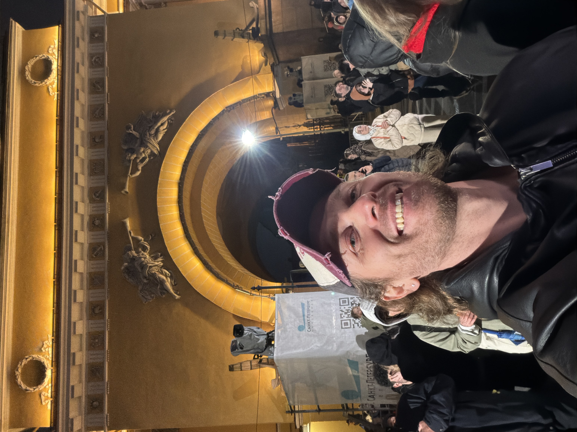 The image shows a man taking a selfie in the Winter Palace Square in Saint Petersburg, Russia. He is wearing a baseball cap and a black leather jacket. Behind him is the General Staff Building, featuring ornate architectural details, including sculptures and arches. A crowd of people is gathered in the square. There is a booth with signage in Russian, including a QR code and the name "Санкт-Петербург" (Saint Petersburg). A television camera is visible on a tripod near the booth. The scene is illuminated with artificial light, suggesting it is evening or nighttime.