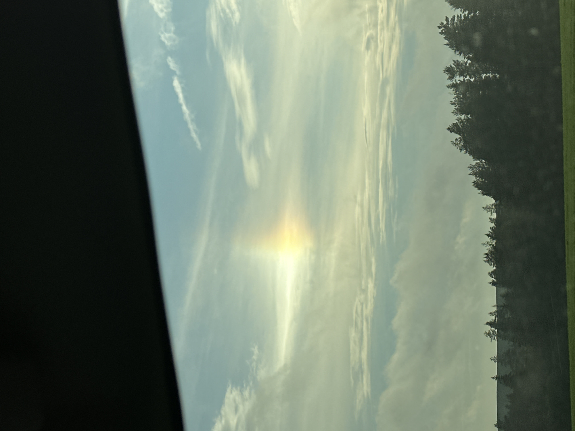 The image shows a sky with wispy clouds and a sun pillar. A dark, curved object, possibly a car roof or visor, occupies the top of the frame. Below the sky is a dark line of evergreen trees silhouetted against the horizon, with a green field in the foreground. The scene is likely a landscape view from inside a vehicle. There is no indication of Buranovo, Russia, or any specific location within it.