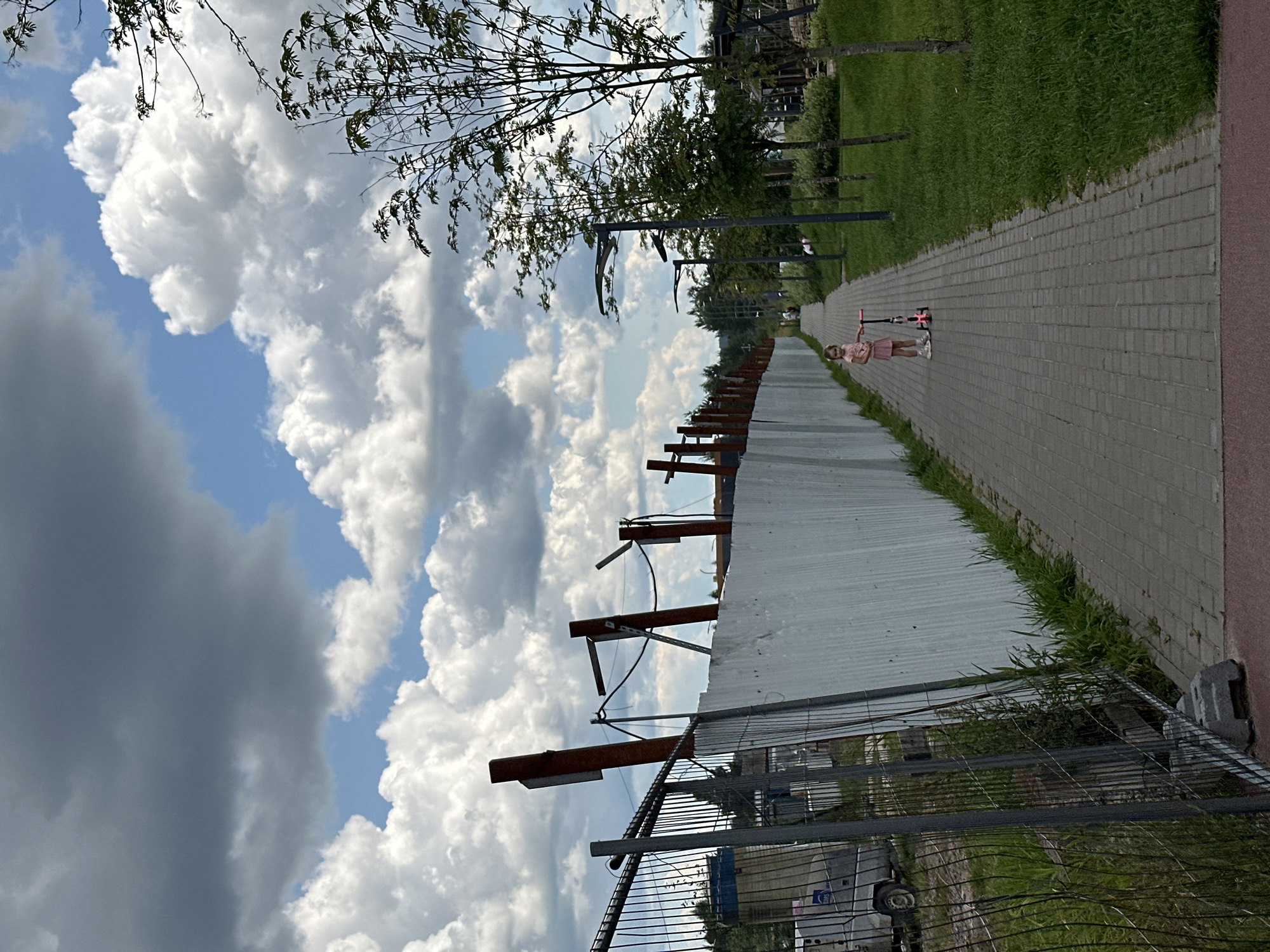 The image depicts a paved pathway in an urban park area. On the left, a temporary construction fence, made of metal mesh and white panels, runs along the pathway. Beyond the fence, there is evidence of construction, including a vehicle and some building materials. A young girl, dressed in a pink outfit, stands on the pathway with a pink scooter. She is facing away from the viewer, looking towards the end of the pathway. The pathway is lined with green grass and trees on the right side, with modern-style lampposts interspersed. The sky above is filled with dramatic clouds, some dark and grey, others bright white against a blue sky. The scene appears to be in a park or recreational area, possibly undergoing development or renovation. This image was taken on August 3, 2023, at 1:34 PM. The location is Vsevolozhsk, Russia.