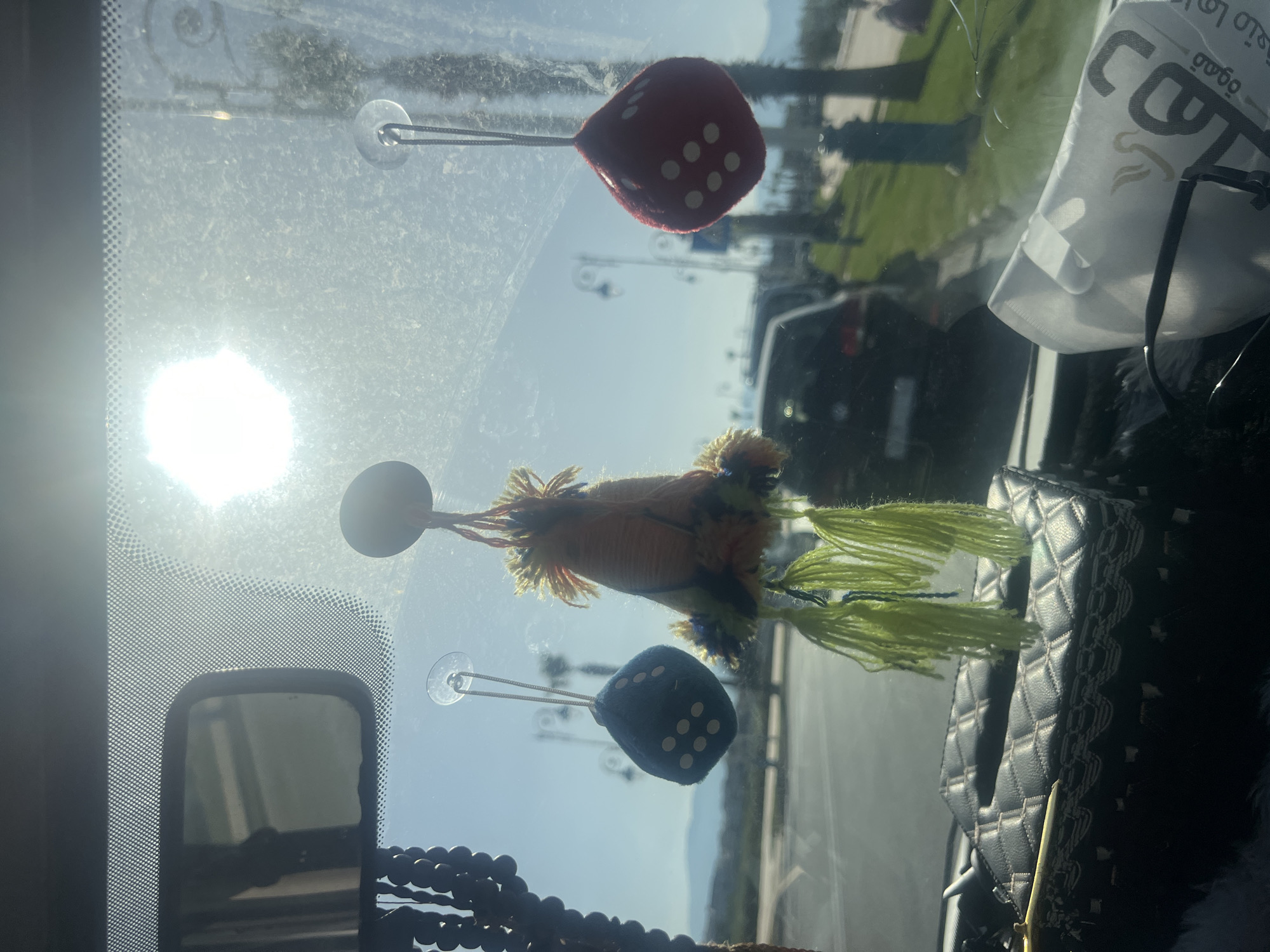 The image shows the interior of a vehicle, likely a car, with the dashboard and windshield partially visible. Hanging from the windshield via suction cups are decorative items: a red fuzzy dice, an object with orange and yellow tassels, and a blue fuzzy dice. A string of beads is partially visible near the rearview mirror. A black quilted bag or purse sits on the dashboard. Through the windshield, a street scene is visible, featuring greenery, streetlights, and other vehicles. The location is identified as Martil ⵎⴰⵔْتِيل مرتيل, Morocco. The sun is shining brightly, causing glare on the windshield.