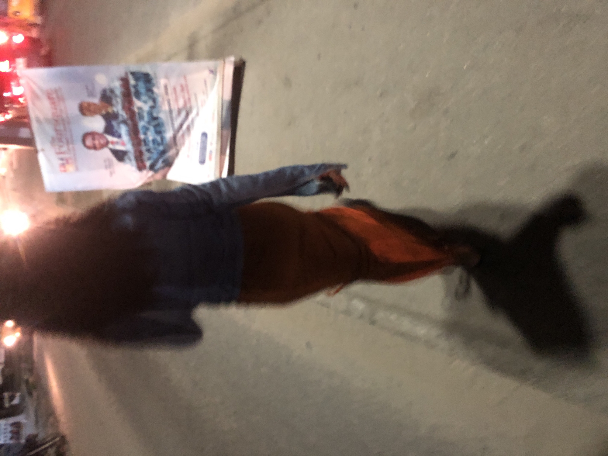 In Ado, Nigeria, a person walks past a large banner on a street at night. The person, seen from behind, wears a blue jacket and an orange skirt. The banner, partially obscured, displays images of two men in suits, likely political figures, with text in English. The street is paved and appears to be well-lit by artificial sources, casting shadows. The environment suggests an urban setting with some visible vehicles and streetlights in the background. The image is somewhat blurred, indicating motion or low light conditions.