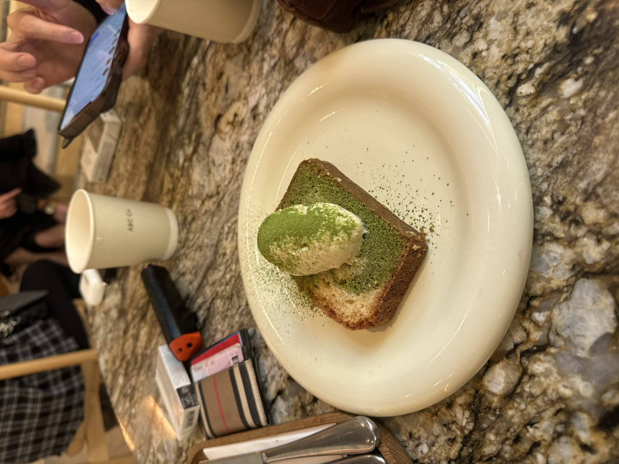 The image shows a white plate with a slice of matcha cake topped with a scoop of cream or ice cream and sprinkled with matcha powder. The plate sits on a table with a marble or granite-like surface. Next to the plate, there is a wallet with a Burberry-like pattern, a black object, and a cup. In the background, a person is holding a phone and there are other people and objects. The location in the city of Moscow, Russia cannot be determined from the image.