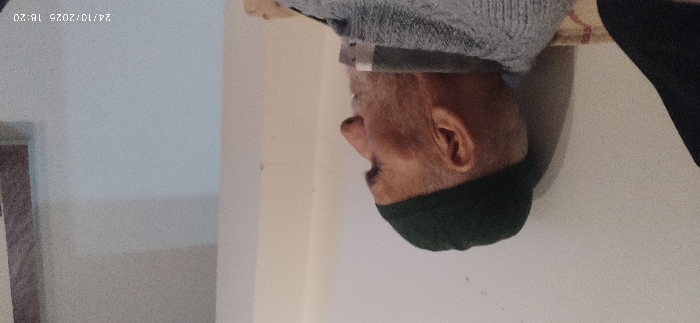 The image, captured on 24/10/2025 at 18:20, displays an elderly male seated indoors in Mu'adammiyat ash-Sham, Syria. The individual is presented in profile, facing right, wearing a dark green skullcap and a grey, possibly knitted, long-sleeved garment over a white-collared undershirt. He has a short, white beard. The immediate background is a light-colored, plain wall, with a partial wooden frame visible on the far right. A patterned fabric, indicative of upholstery or a cushion, is visible beneath the subject. A dark, indistinct object is partially visible at the far left of the frame.