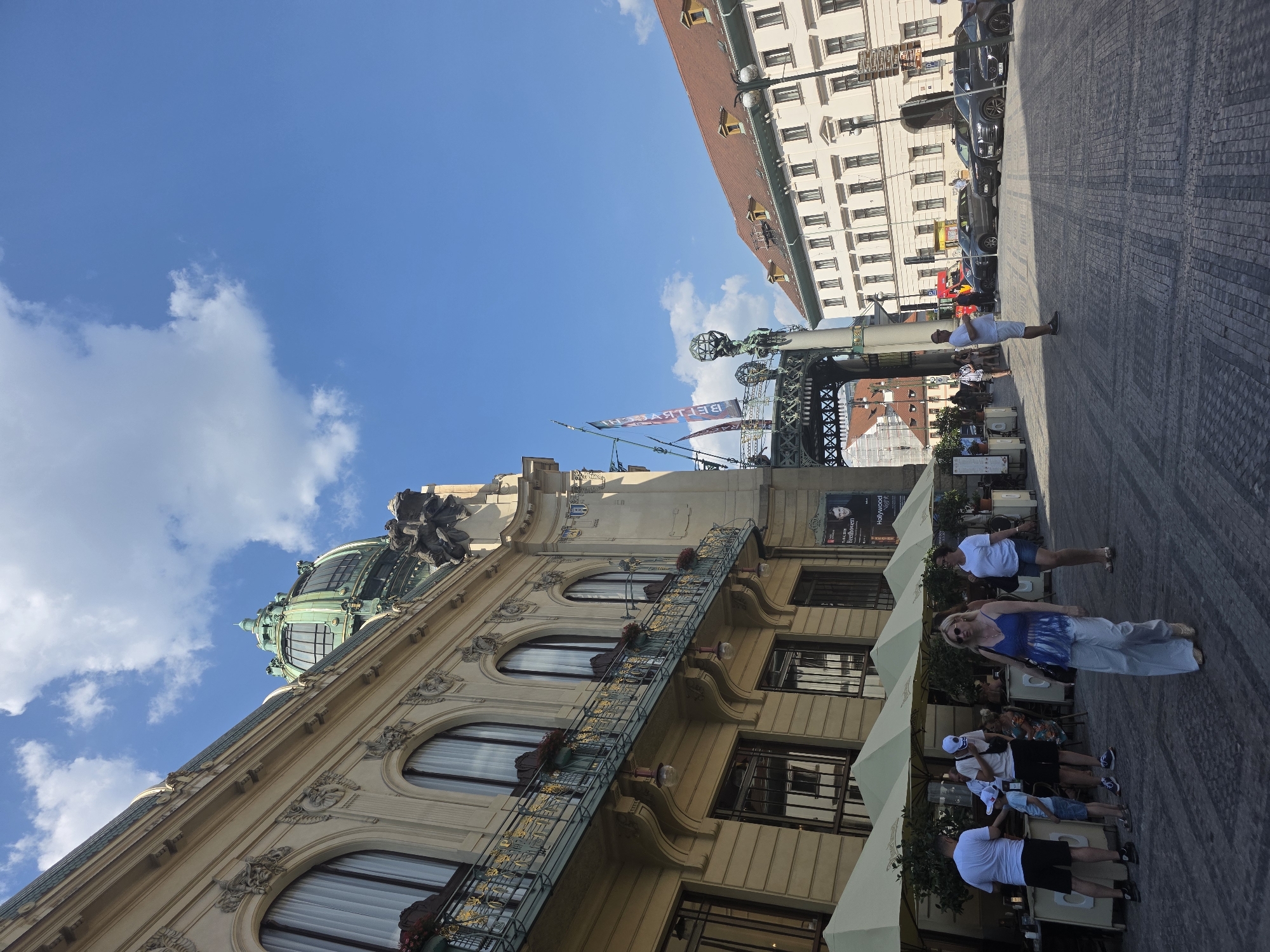 This is a low-angle, wide shot of a bustling street scene under a bright, sunny sky with scattered white clouds. The image is rotated 90 degrees counterclockwise.
In the foreground, a woman in a blue and white patterned top and light-colored pants walks towards the viewer. Behind her, a group of people are seated at outdoor tables under large, cream-colored umbrellas, suggesting a cafe or restaurant. Further back, a man in a white t-shirt and shorts stands on the cobblestone pavement, looking towards the right. Several parked cars are visible along the side of the street.
The background features impressive architecture. On the left, a light-colored building with ornate decorative elements and arched windows is prominent, topped by a green dome. To the right, a taller, cream-colored building with a red-tiled roof extends upwards, also featuring numerous windows. Banners with red and blue lettering hang from a metal structure between the buildings. One banner clearly reads "BETLSASCIN" (reversed). Another sign higher up on a building on the left shows a black and white image of a man and the text "HOLLYWOOD".
The scene appears to be a lively urban environment, likely during the daytime due to the strong sunlight and shadows. The weather seems pleasant, with clear skies. The overall mood is one of everyday activity and urban exploration.