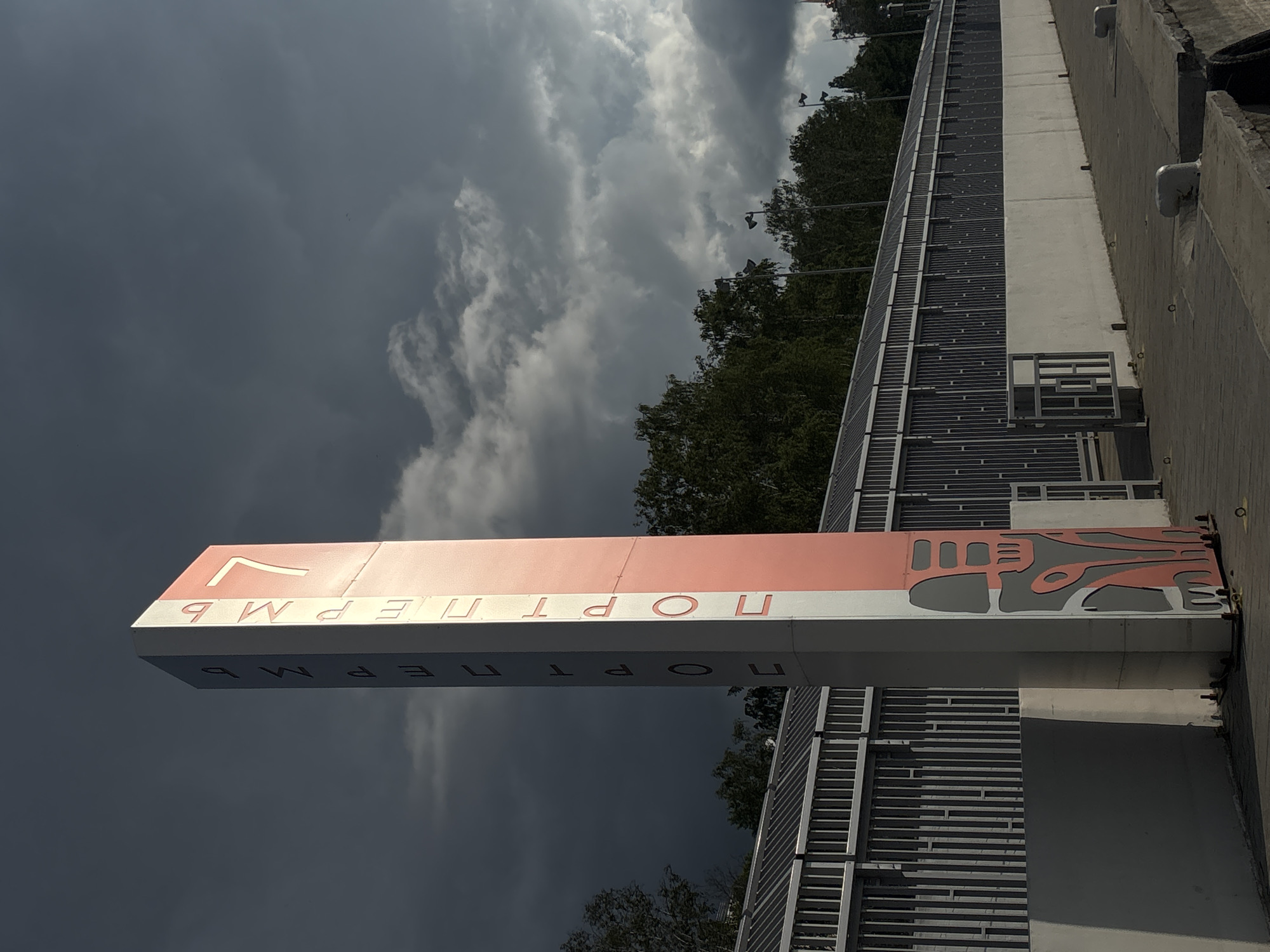 This image depicts a tall, rectangular, multi-sided pillar serving as a sign, prominently featuring the text "ПОРТ ПЕРМЬ" (Port Perm) in Cyrillic vertically on at least two of its visible faces. The pillar's surfaces are predominantly white or light grey, with one visible side displaying a reddish-orange or salmon color. An abstract, stylized design, composed of cut-out shapes in grey and reddish-orange, is present on the lower section of the pillar's colored face. The pillar stands on a concrete paved area, which appears to be a dock or waterfront promenade.
In the background, a long, continuous structure with a corrugated metal roof and vertical slats or panels extends horizontally across the scene. This structure is fronted by a low, light-colored concrete wall. Dense green foliage and trees are visible above and behind this structure, extending towards the horizon. The sky is heavily overcast with dark, ominous clouds dominating the upper portion of the frame, though some lighter grey clouds are also visible, particularly towards the right. The pillar is brightly illuminated, creating a strong contrast against the dark sky.
The concrete paved area in the foreground includes several small, white or light grey mooring bollards or cleats, indicating its function as a port or harbor. A section of a metal fence or gate is visible to the left of the pillar, and another small metal gate is partially visible further along the concrete wall to the right. No individuals are present in the image. The scene is located at the Port of Perm, Russia, as indicated by the sign.
