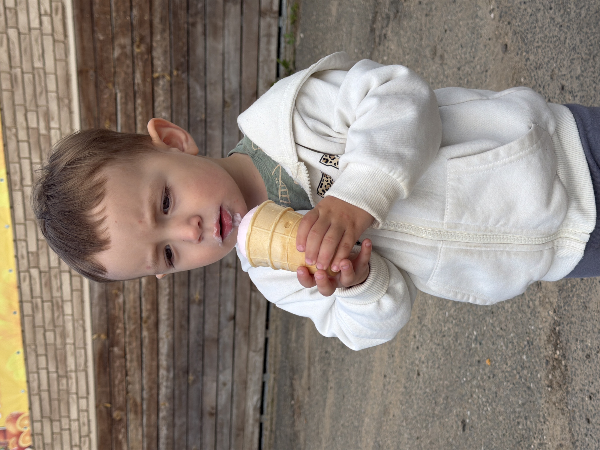 A young child, likely a toddler, stands outdoors, holding a pink ice cream cone in a waffle cone. The child wears a white hooded zip-up jacket over a green t-shirt and grey pants. There is a smudge of ice cream on the child's upper lip and chin. The background consists of a wooden wall and a brick wall, suggesting an outdoor seating area or a public space. The ground is paved with asphalt and small stones. The lighting indicates it is daytime. The city of Красный Октябрь, Russia, is mentioned in the prompt, but the image itself does not contain any specific visual cues that place it in that particular location.