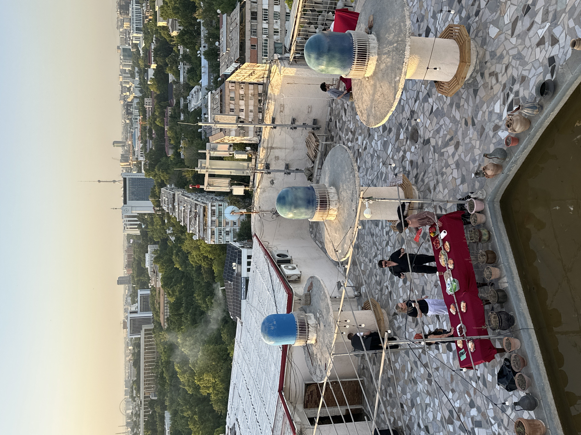 Captured from an elevated perspective, the image shows a rooftop space in Tashkent, Uzbekistan. The roof is adorned with a mosaic tile pattern and lined with decorative pots along the perimeter. Distinctive blue-domed structures are intermittently placed on the rooftop. A long red table is set up, displaying food items, with several people gathered around it. The backdrop reveals the city skyline with a mix of buildings and green spaces, including a prominent television tower and what seems to be a Ferris wheel in the distance.
