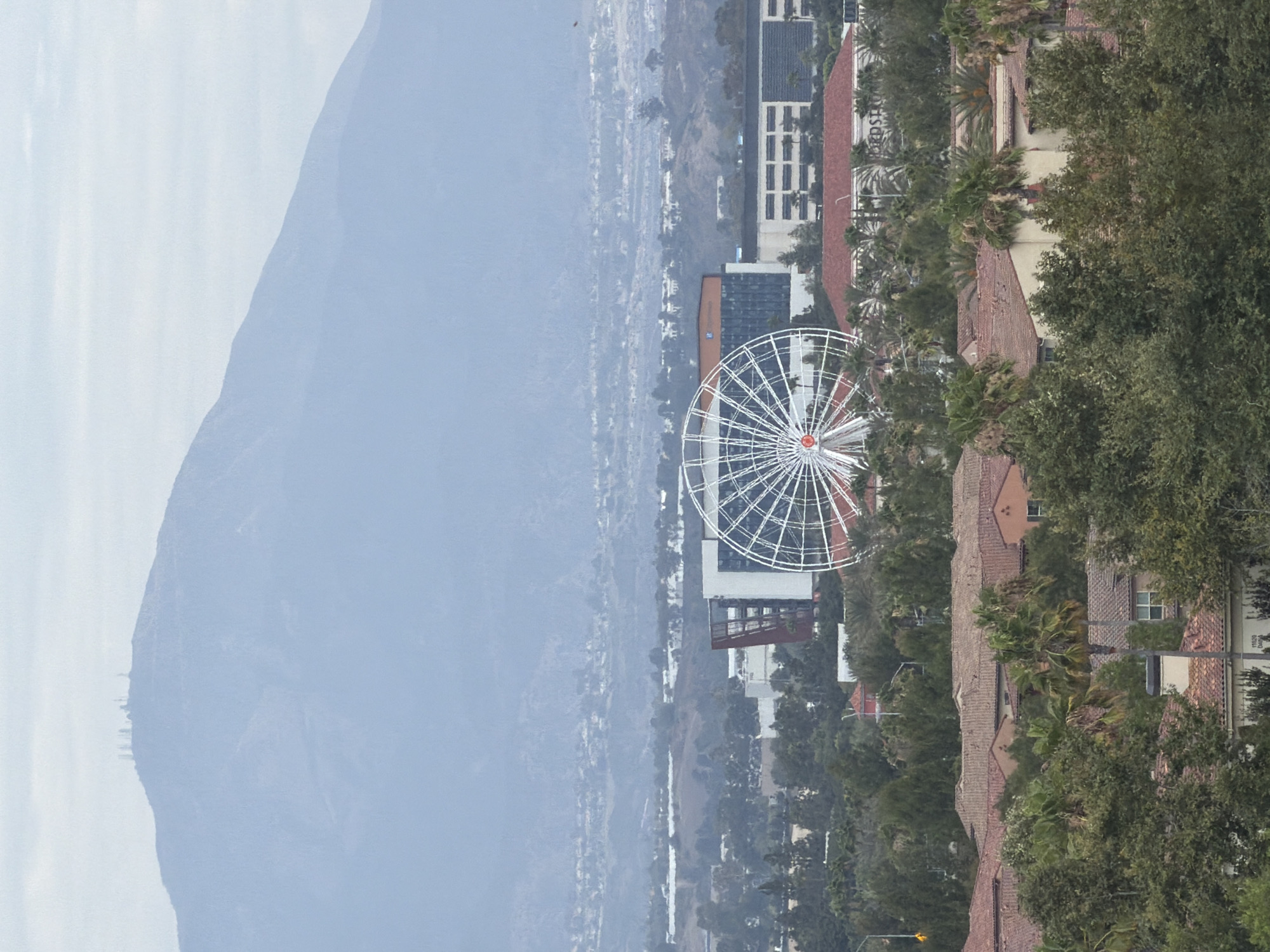 This image, captured in Irvine, California, presents a panoramic view of a cityscape nestled against a backdrop of hazy mountains. The foreground is dominated by a large, white Ferris wheel, its intricate spokes forming a striking geometric pattern against the muted sky. Several buildings with red-tiled roofs are visible, interspersed with lush green trees, including palm trees, suggesting a warm climate.
In the distance, a sprawling urban landscape stretches out, fading into a soft, atmospheric haze. The mountains in the far background appear muted and soft, their details obscured by the atmospheric conditions. The overall impression is one of a serene, perhaps overcast day. The presence of the Ferris wheel suggests a location that might be an entertainment district or a part of a larger complex. The architectural style of the buildings, with their red roofs and white facades, is typical of Southern California. No people are visible in the frame.
The image is oriented vertically, which may imply it was taken with a phone camera or has been rotated. The lighting suggests it is daytime, but the heavy haze or fog softens the light and obscures finer details, creating a diffused and tranquil atmosphere. The scene conveys a sense of scale, contrasting the man-made structures with the natural landscape.