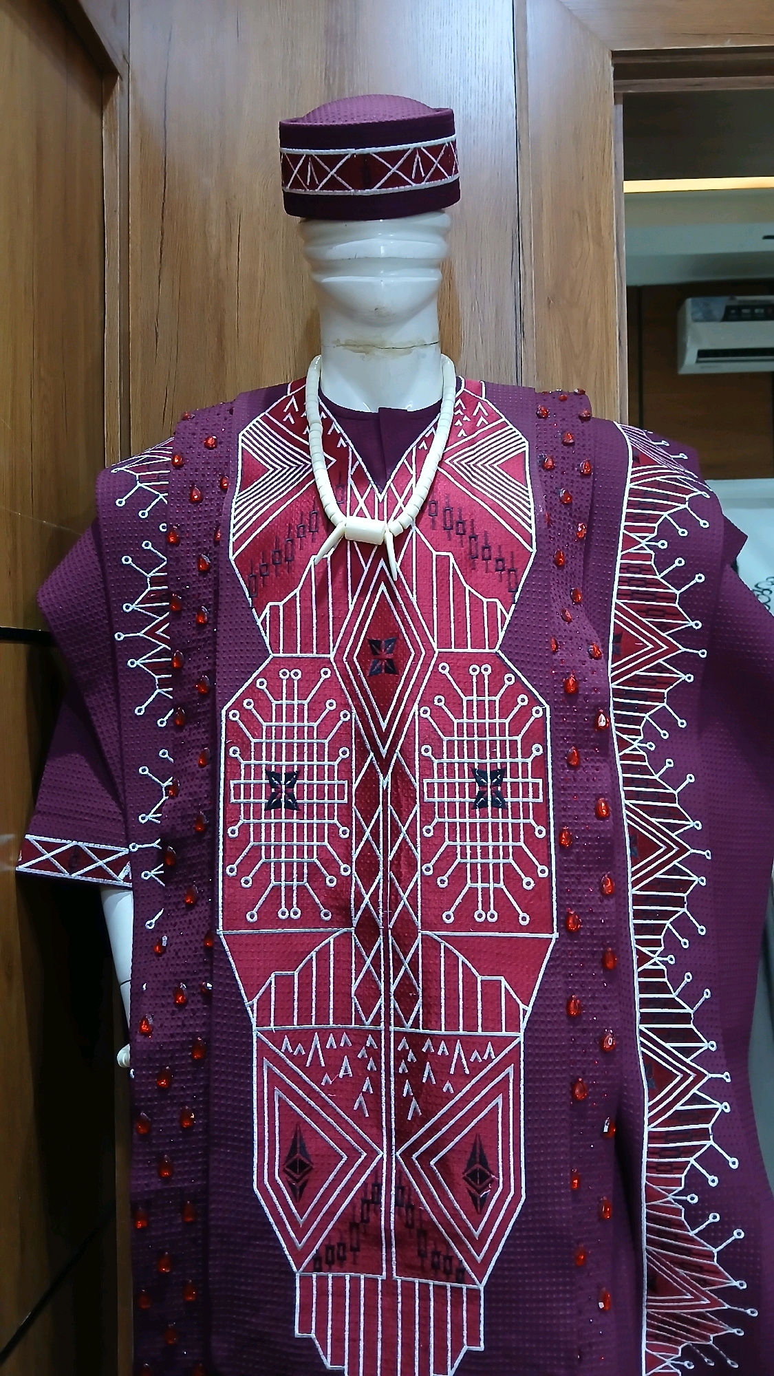 An ornate, burgundy traditional African garment is displayed on a white mannequin. The garment features intricate white geometric patterns and is adorned with small, red jeweled accents. A matching burgundy cap with white trim sits atop the mannequin's head, and a necklace made of ivory-colored beads and two pointed tusks rests around its neck. The mannequin is positioned against a backdrop of warm-toned wooden paneling.