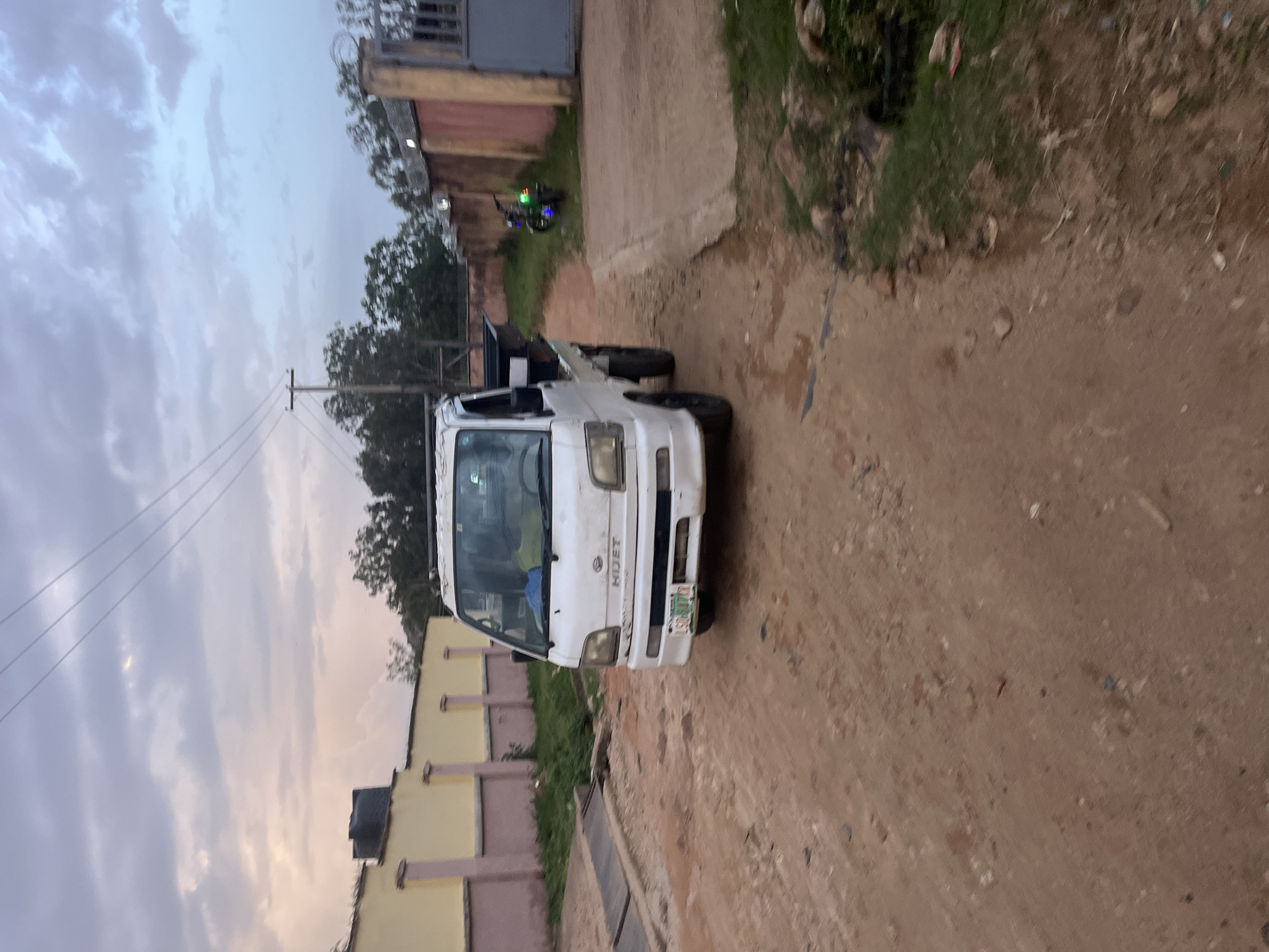 The image shows a white Daihatsu Hijet truck parked on a dirt road. The truck has a black bed and appears to be slightly dirty. A license plate with the letters "LSG 817 ZK" is visible on the front of the truck. In the background, there are buildings with yellow and pink walls, trees, and a motorcycle with green lights. The sky is cloudy with hints of pink and blue, suggesting it might be dusk or dawn. The scene is likely located in Gura Top, Nigeria, as indicated. The environment appears to be somewhat rural or semi-urban, with unpaved roads and vegetation.