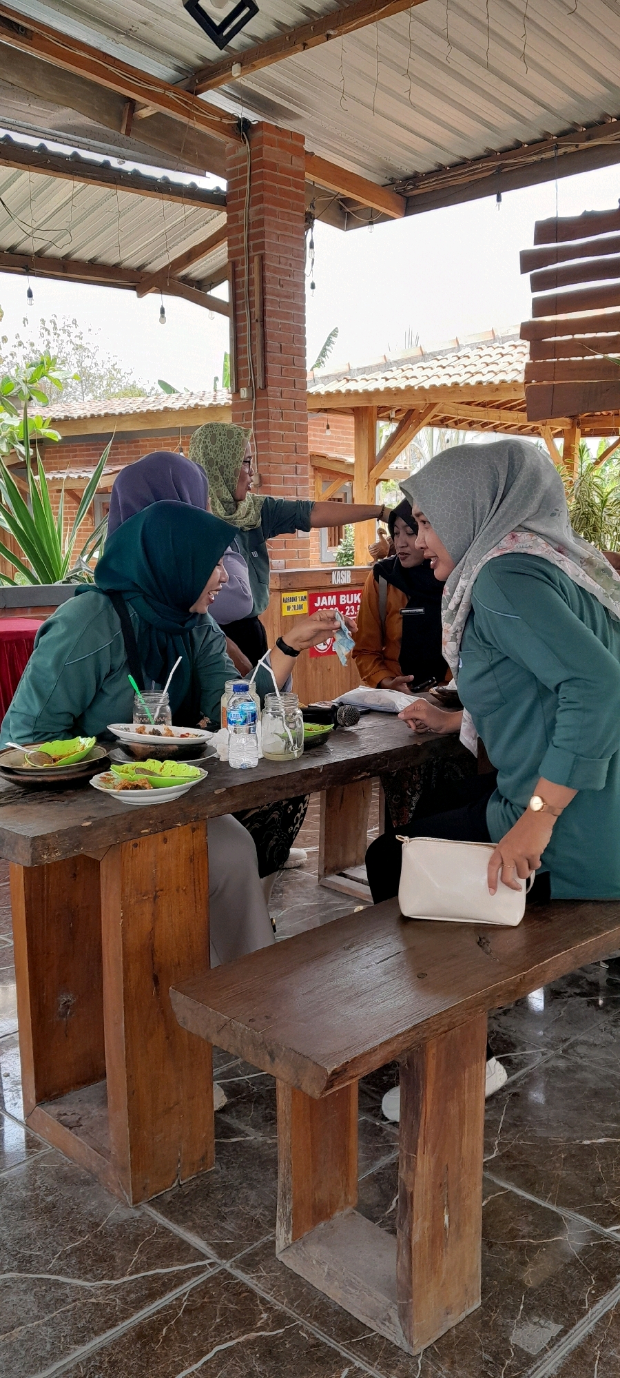 In Sidohasri, Indonesia, at an outdoor dining establishment with a rustic, covered seating area, four women are gathered around a wooden table. The setting appears casual, possibly a warung or restaurant. The women, all wearing hijabs, are engaged in conversation and eating.
In the foreground, two women are seated at the table. The woman on the left, wearing a teal hijab and jacket, is smiling and holding a piece of paper money, possibly making a payment or exchange. She has a plate with food and a drink in front of her. To her right, a woman in a grey hijab and teal jacket is leaning forward, gesturing with her left hand and speaking to the woman on the left. A white clutch bag rests on the wooden bench next to her.
In the background, two other women are partially visible. One, wearing a light green hijab and dark jacket, is standing and pointing to her right, appearing to direct attention or give instructions. Another woman, in an orange top and black hijab, is seated behind the table, looking towards the woman in the green hijab.
The table is laden with plates of food, glasses, a bottle of water, and a microphone. The floor is tiled with a marble-like pattern. The structure above is made of corrugated metal roofing supported by wooden beams and a brick pillar, suggesting an open-air or semi-open structure typical of Indonesian outdoor eateries. Lush green foliage is visible in the background, indicating a tropical environment. The overall atmosphere suggests a social gathering with a meal.