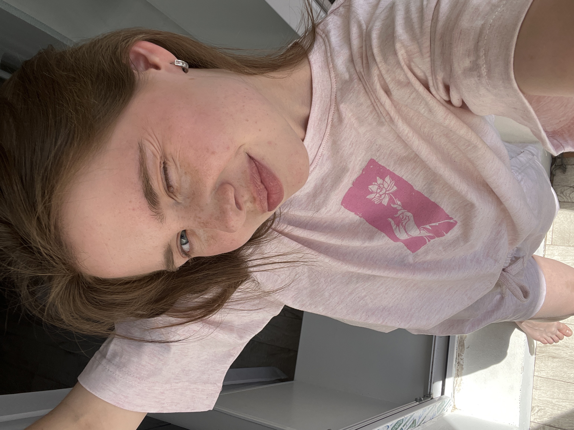 A young woman with auburn hair and freckles is taking a selfie. She is wearing a light pink t-shirt with a pink graphic of a hand holding a lotus flower. She is winking and smiling at the camera. Her right arm is raised and her hand is resting on her head. She is wearing a small silver hoop earring in her right ear. The background appears to be an indoor setting with bright natural light coming from a doorway. The flooring is wood-toned. The overall impression is casual and lighthearted. The provided crops indicate that the image was likely taken in a domestic environment in Chaikovsky, Russia.