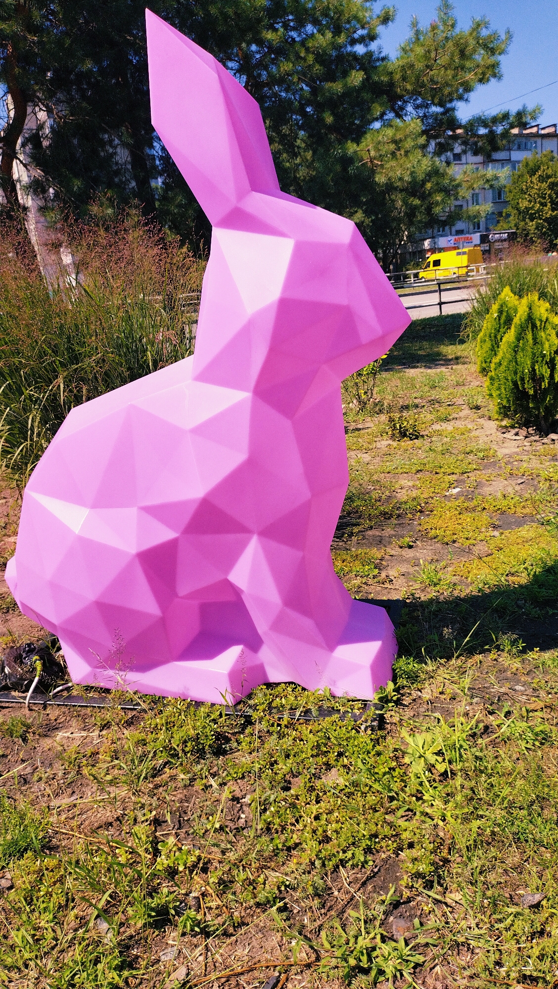 A large, polygonal, light pink sculpture of a rabbit sits in a patch of grass. The rabbit is facing right, with its head tilted slightly upwards. Behind the rabbit, there is a road with a yellow truck driving on it, and a building in the background. Trees and bushes surround the scene. The sky is clear and blue. The image was taken in Horishni Plavni, Ukraine.