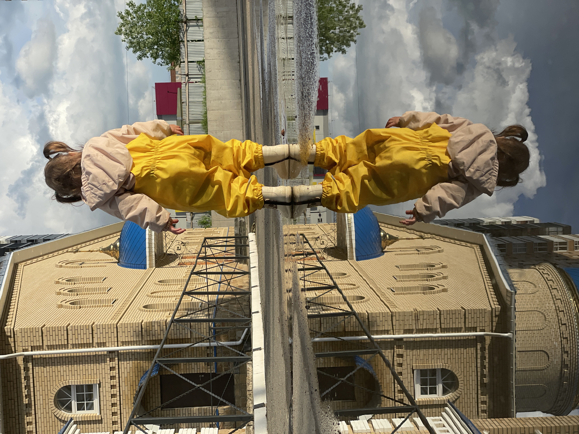 The image depicts a reflection scene, likely in Izhevsk, Russia, given the distinct architectural style of the Russian Orthodox church visible in the lower half of the image. The top half of the image shows a reflection of a young girl in yellow pants and a beige jacket. She has her hair in pigtails. The background of her reflection is a cloudy sky, greenery, and a building with a red roof. The lower half of the image shows the physical structure of the church, likely brick, featuring a blue dome and decorative architectural elements. A metal ladder is visible against the church's exterior. The reflection creates a symmetrical effect, with the child and the church seemingly connected at a central point. There are elements of water visible between the reflection of the girl and church, suggesting the reflections are of a body of water, perhaps a fountain.