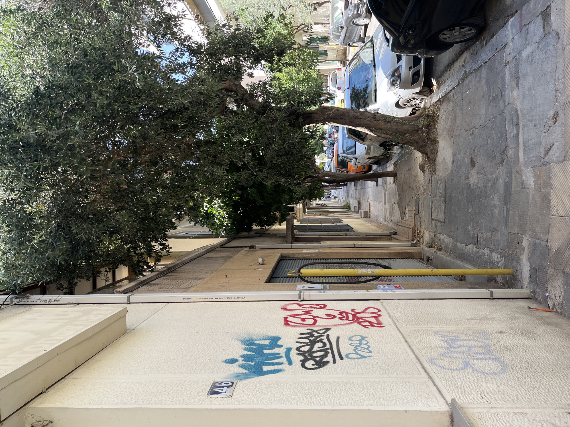 The image depicts a street scene in Athens, Greece, likely on a sidewalk. In the foreground, a textured, light-colored wall of a building features graffiti. To the left, a blue house number "46" is visible. The graffiti includes blue, black, and red spray paint tags, some resembling abstract shapes and figures. A yellow pole with a black loop attached is positioned vertically against the building wall further down.
The sidewalk itself is paved with stone slabs, some of which are cracked and uneven, with dirt and debris scattered in places. Several trees with dense green foliage are planted along the sidewalk, their roots encroaching on the paving.
Beyond the sidewalk, parked cars are lined up along the street. These include a dark SUV on the right, a silver hatchback in the center, and other vehicles including an orange car further down the street. The buildings lining the street appear to be residential or commercial, with windows and balconies visible. The overall impression is of a typical urban street in Athens on a sunny day.
