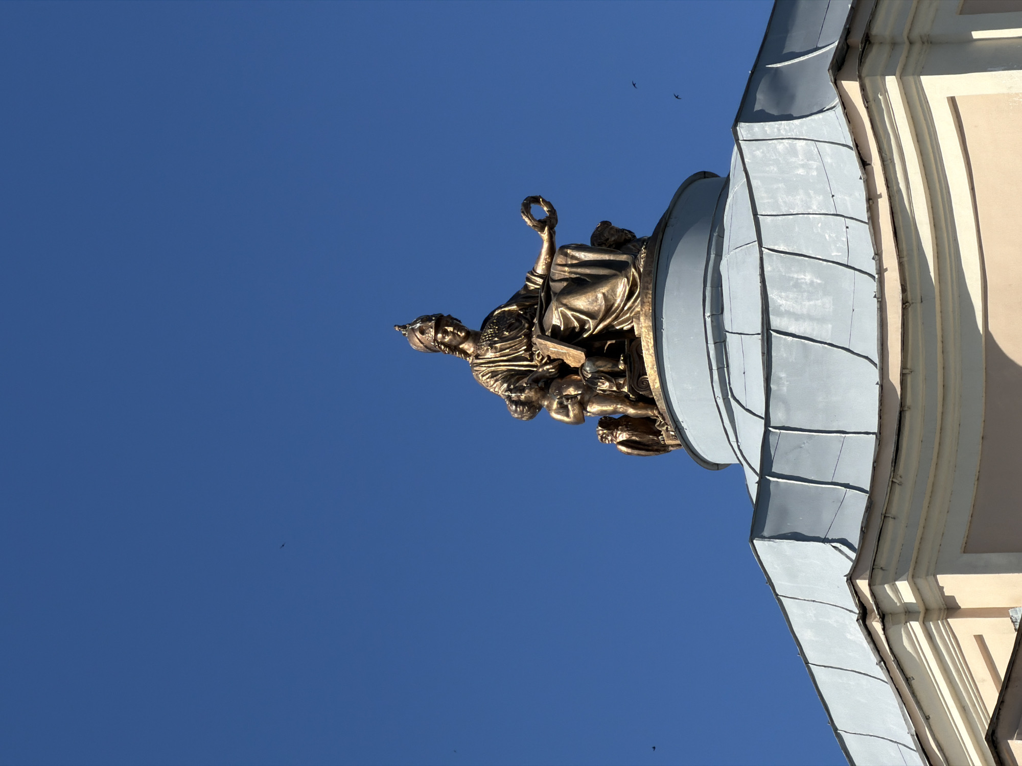 An upward-angled shot captures a bronze allegorical statue adorning a grand classical building under a clear, deep blue sky. The central figure of the sculpture is a seated female, possibly a goddess or personification, wearing a helmet or crown and holding a laurel wreath or crown aloft. Smaller, infant-like figures or putti are visible around her base, contributing to the ornate sculptural group.
The statue is positioned on a curved architectural element, likely a dome or cupola, clad in distinct segmented gray metal panels that reflect the sunlight. To the right, a section of the building's facade is visible, characterized by light-colored, smooth stone or stucco walls with vertical fluting or pilaster-like detailing. Bright, direct sunlight illuminates the sculpture and the building, casting crisp shadows that define the architectural forms and suggest a clear, sunny day, likely mid-morning to afternoon.
Several small, dark bird silhouettes are scattered across the vast expanse of the blue sky, adding a sense of scale and movement. The scene is devoid of any visible text. The architectural style and monumentality are characteristic of structures found in Saint Petersburg, Russia, where the image was taken.