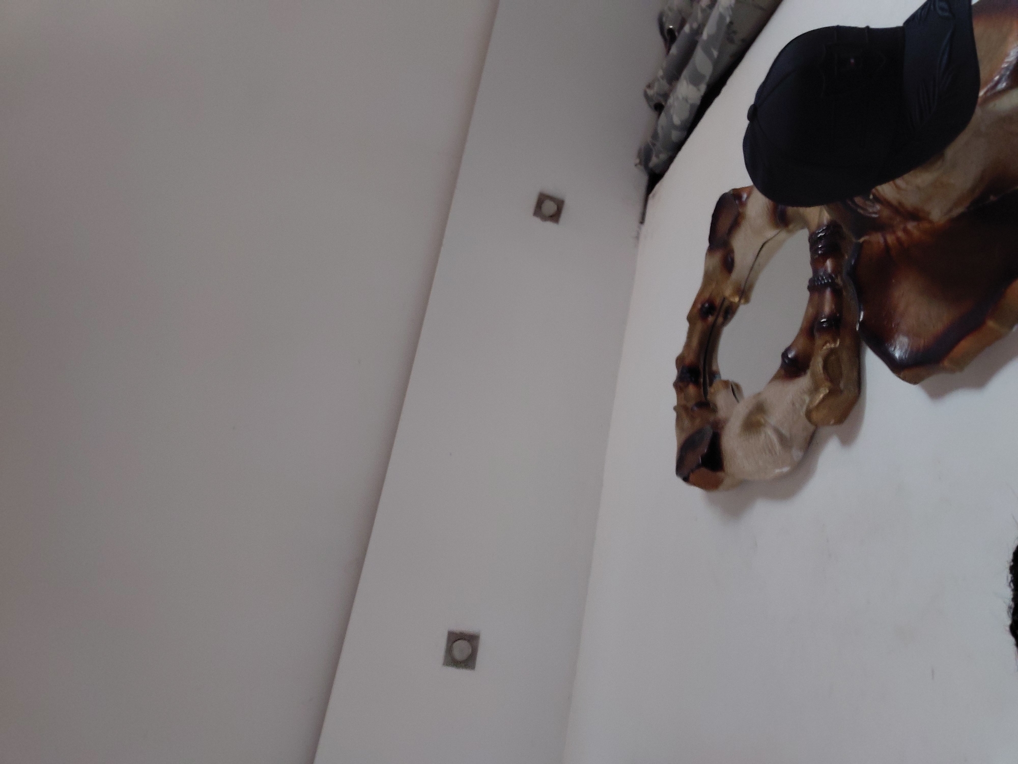 The image presents an indoor scene with a tilted perspective, primarily featuring light-colored, possibly white, walls. On the right side of the frame, a large, organic-shaped decorative object, likely made of wood or a resin, is prominent. It exhibits a blend of dark brown and lighter beige or tan hues, with a somewhat glossy finish, and features an irregular, abstract design that incorporates a small circular mirror.
Resting on top of this decorative piece is a black baseball cap. In the upper right corner, a glimpse of camouflage-patterned material, possibly part of clothing or another item, is visible. The wall extending towards the left and center of the frame features two distinct square-shaped fixtures, each with a circular element in its center, which could be small lights or electrical covers. The lighting is diffused and even, suggesting indoor illumination. There is no activity or visible text in the image. The setting appears to be a static interior space.