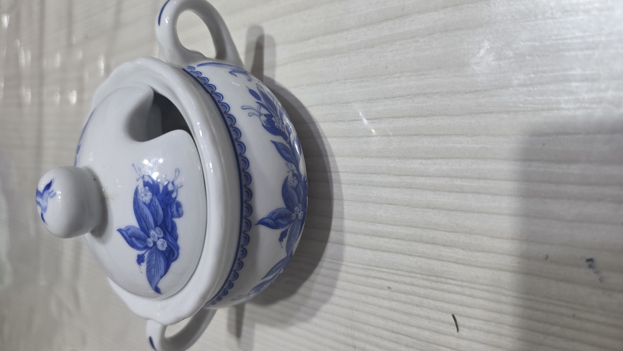 A white ceramic sugar bowl with blue floral patterns sits on a light-colored wooden surface. The sugar bowl has two handles and a lid with a knob. The lid has a cut-out to accommodate a spoon. The background is blurred, but appears to be a similar wooden surface. The scene is indoors, likely on a table. There are no people or animals present. The image is a close-up of the sugar bowl, with the bottom half of the bowl and the wooden surface visible. There are no discernible landmarks or indications of Chuy, Kyrgyzstan.