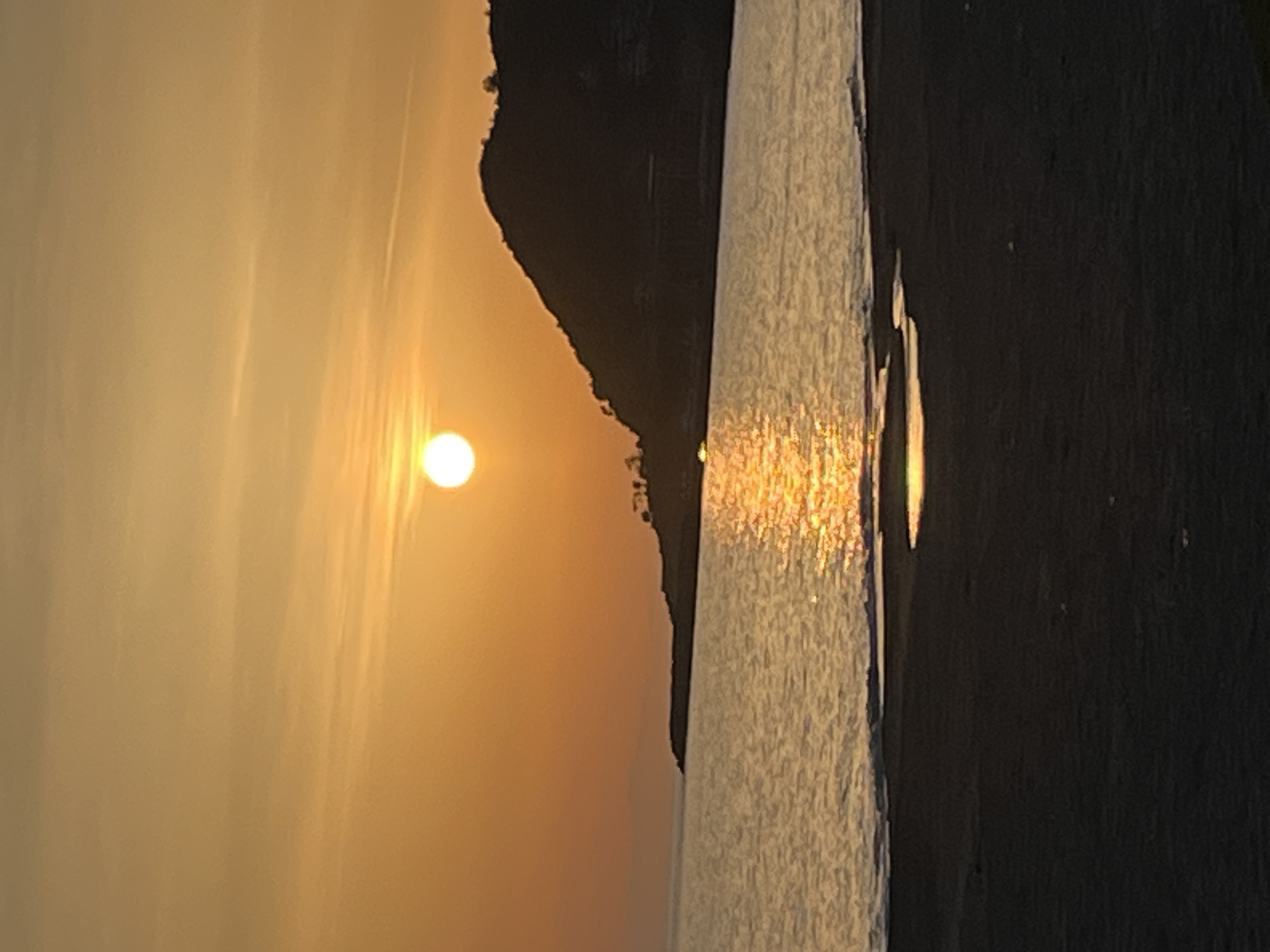 The image depicts a sunset over water, likely a sea or lake, with a dark landmass or headland silhouetted against the sky. The sun is a bright orb low on the horizon, casting a golden glow across the water surface. The water surface is textured with visible reflections of the sunlight. The foreground appears to be a dark, possibly rocky, shoreline. This image is likely taken in Alanya, Turkey given the landscape and sunset.