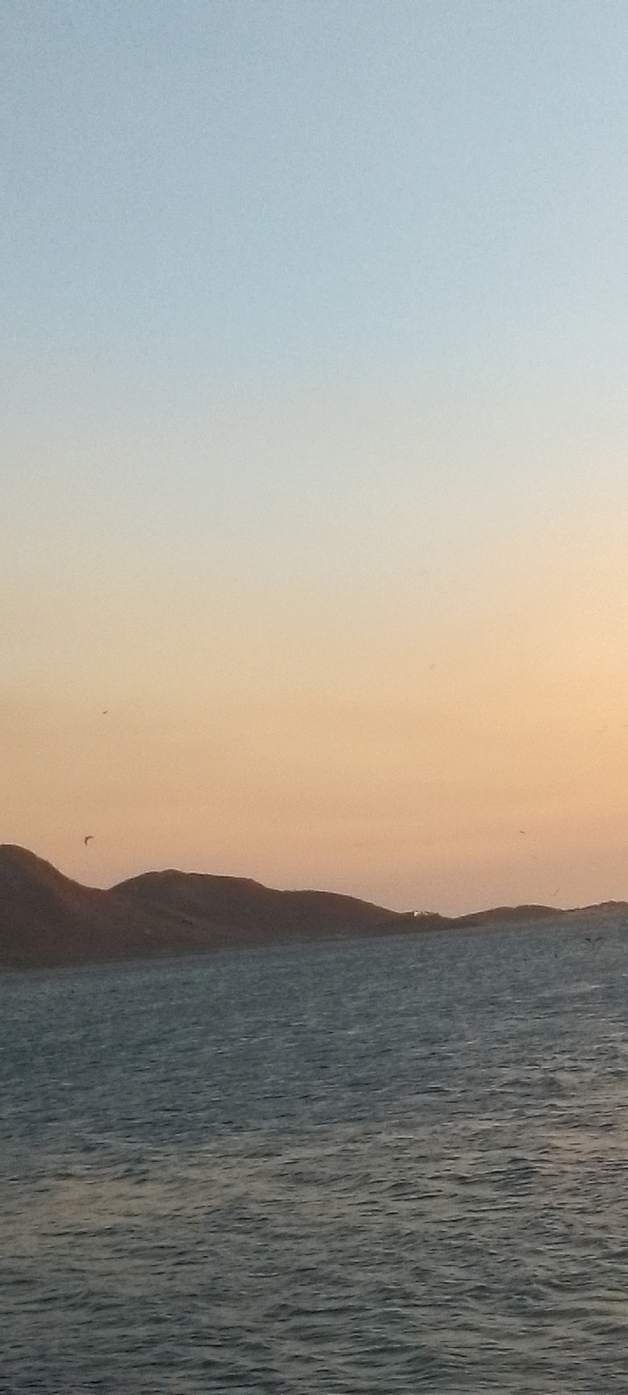 The image shows a view of the Caribbean Sea in Juan Griego, Venezuela, at dusk or dawn. The sky transitions from light blue at the top to a warm orange near the horizon. There are a few indistinct objects (perhaps birds or small watercraft) near the horizon, which would be the Caribbean Sea. Several low-lying hills/mountains are silhouetted against the sky, and these are mostly the Cerro La Vieja on the left, followed by the Cerro Vista Bella and Cerro La Loma with the white fort of La Galera in the center. The sea is calm with gentle waves.