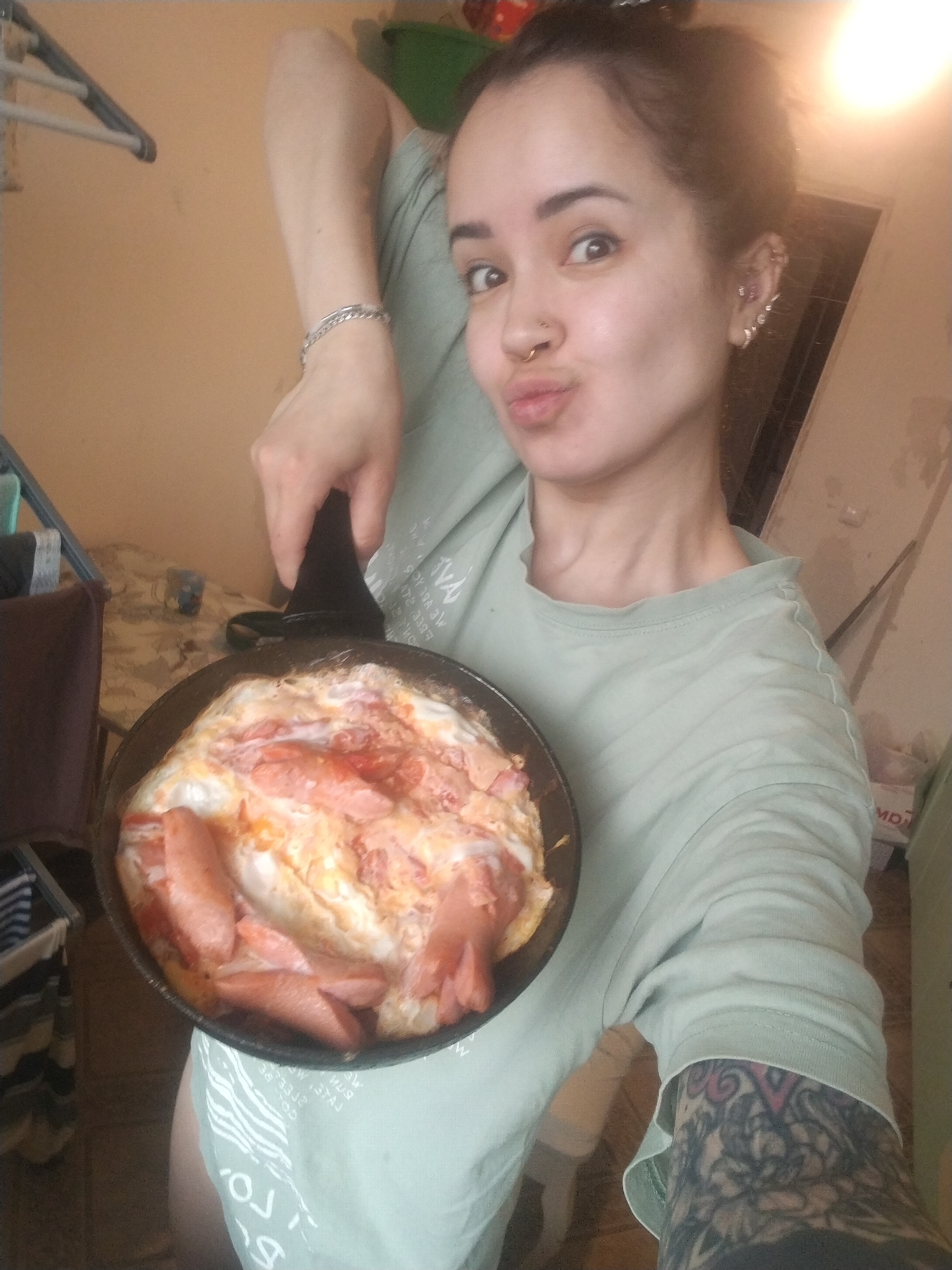 A woman holds a frying pan of cooked eggs and sausages up to the camera. She is light-skinned with dark hair and wearing a light green t-shirt. Her left arm is raised, displaying a silver bracelet and tattoos on her lower arm. She has a nose ring and multiple piercings on her left ear. The background shows a domestic setting with a clothes rack and a table. The city is not determinable from the image.