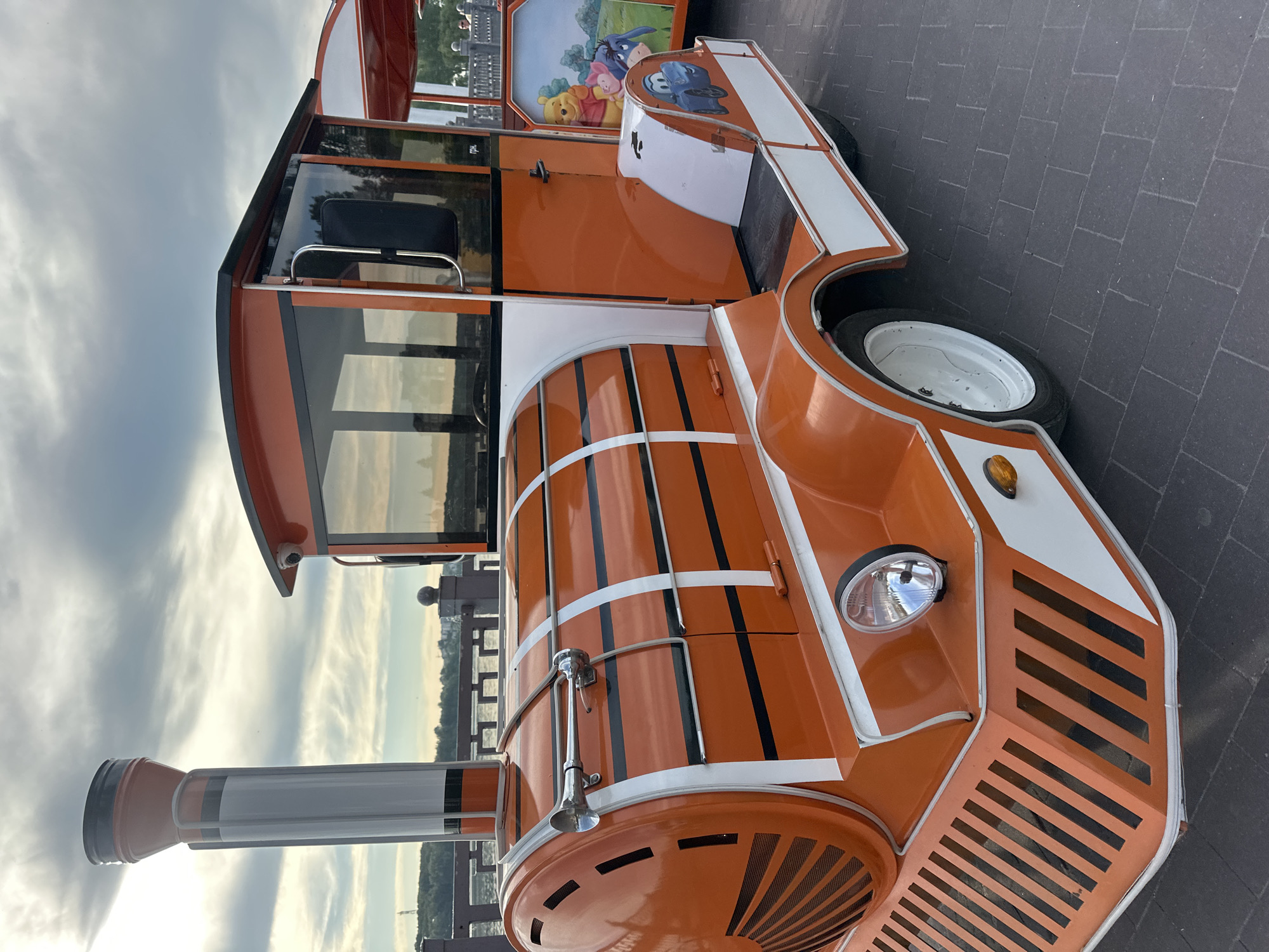 A close-up, ground-level, eye-level, medium shot captures an orange and white toy train on a paved pathway in Ternopil, Ukraine. The train, designed to resemble a tiger, has a prominent smokestack and a circular grille at the front. Black and white stripes adorn its body, and a silver horn is mounted on the side. A single headlight is visible at the front. The train's cabin features large windows, and a side panel displays cartoon characters, including Winnie the Pooh and Eeyore, along with a blue car. In the background, a body of water and a railing suggest a waterfront location. The sky above is partly cloudy with the sun breaking through. The paving stones are dark gray and rectangular, forming a distinct pattern. No people are clearly visible in the immediate vicinity of the train.