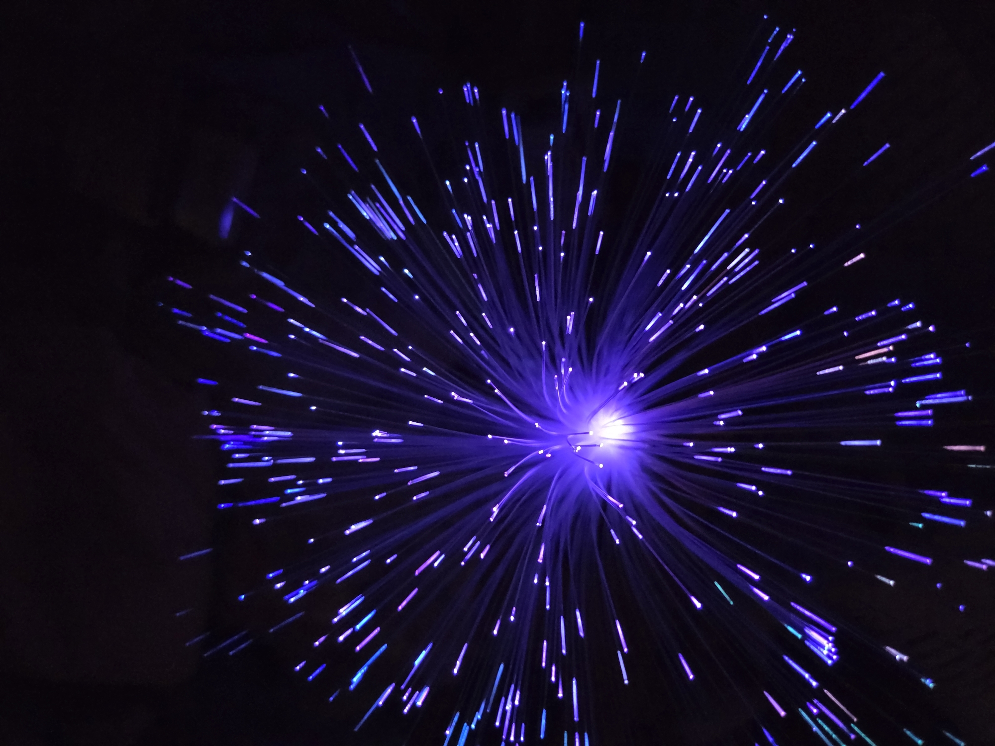 A close-up image shows a fiber optic lamp against a dark background. The light source is concentrated in the center, emitting numerous strands of light that radiate outwards. The strands are primarily blue and purple, with some hints of pink and green. The image lacks identifiable features linking it to Cherkovitsa, Bulgaria, or any specific location. There are no people or actions visible in the image.