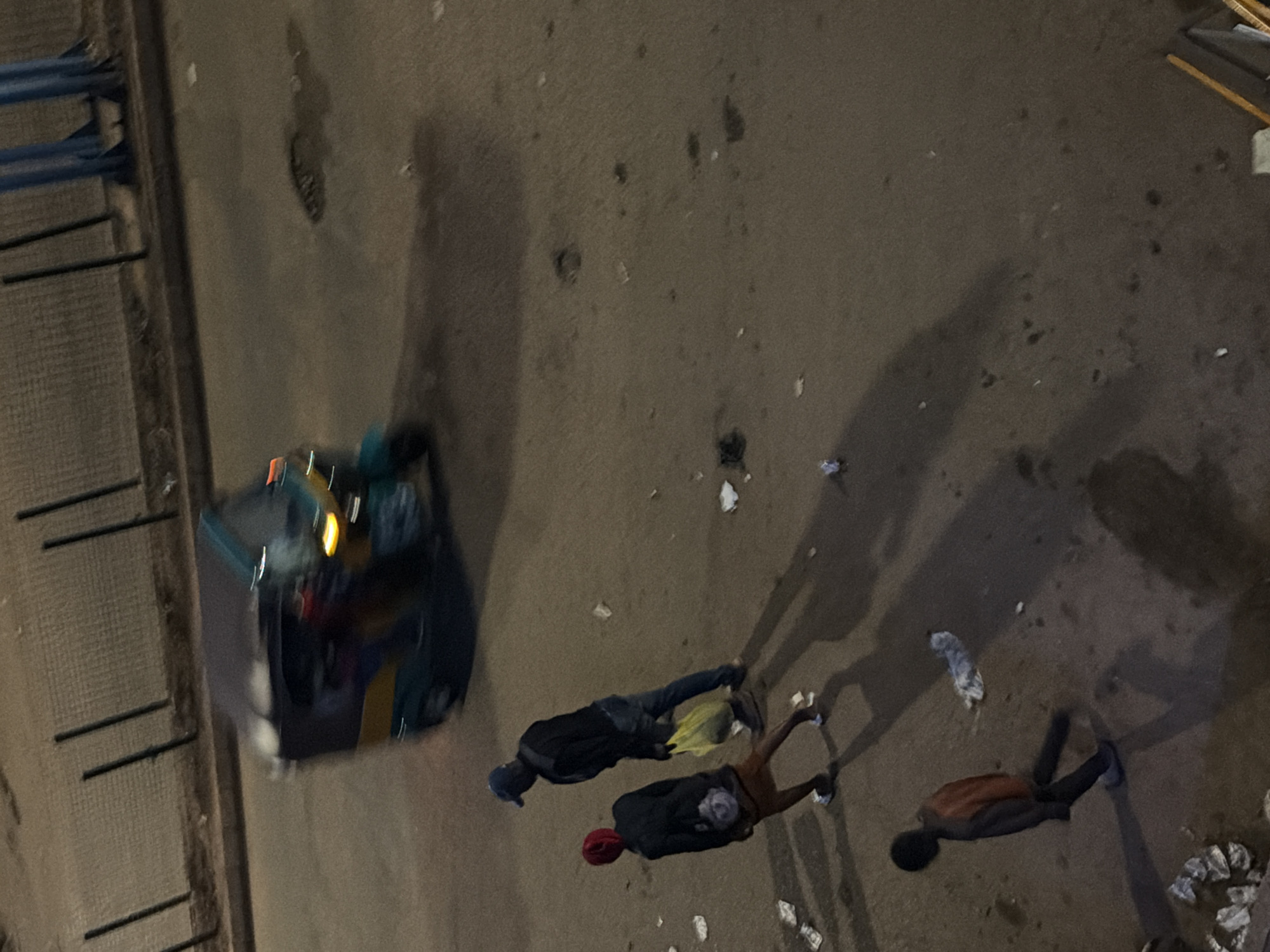 In Bukuru, Nigeria, a blurry image captures a street scene at night. A three-wheeled auto-rickshaw is moving from left to right, its lights faintly illuminated. Two individuals are walking on the asphalt road. The person in front is wearing a red head covering and dark clothing, with a light-colored bag over their shoulder. Behind them, another person, wearing a blue cap and dark attire, carries a yellow plastic bag. A third individual, a child wearing an orange top and dark pants, is walking away from the viewer, casting a long shadow. Scattered across the ground are small pieces of debris and what appear to be discarded papers. The overall impression is of a dimly lit, somewhat unkempt urban environment.