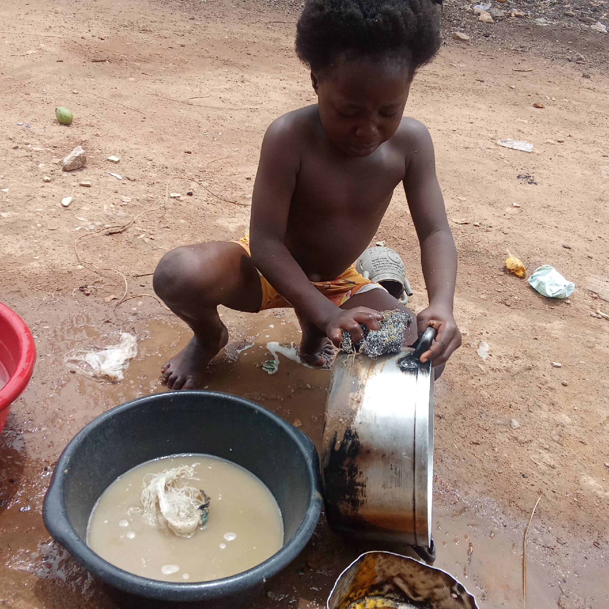 A young child, with dark skin and short, curly dark hair, is depicted squatting on a dirt ground, intently focused on scrubbing a dirty silver pot. The child is shirtless and wears yellow shorts or underwear. They are using a metallic scrubber on the side of the pot, which has visible burnt residue.
Water from the washing process creates muddy puddles around the child's bare feet. A black plastic basin, positioned in front of the child, contains murky, foamy water and some white fibrous material, suggesting it's the wash basin. Partially visible to the left is a red plastic basin, likely holding rinse water.
The outdoor setting is characterized by reddish-brown dirt and mud, with scattered small stones, gravel, and various pieces of debris, including a green fruit (possibly a mango), white plastic, and string. A light-colored plastic container is visible behind the child's right leg. The scene takes place in natural daylight. Faint embossed text on the side of the silver pot reads "D 30 cm" and "IND" (possibly indicating "INDIA"). The image portrays a child performing daily chores in what appears to be a rural or less developed area.