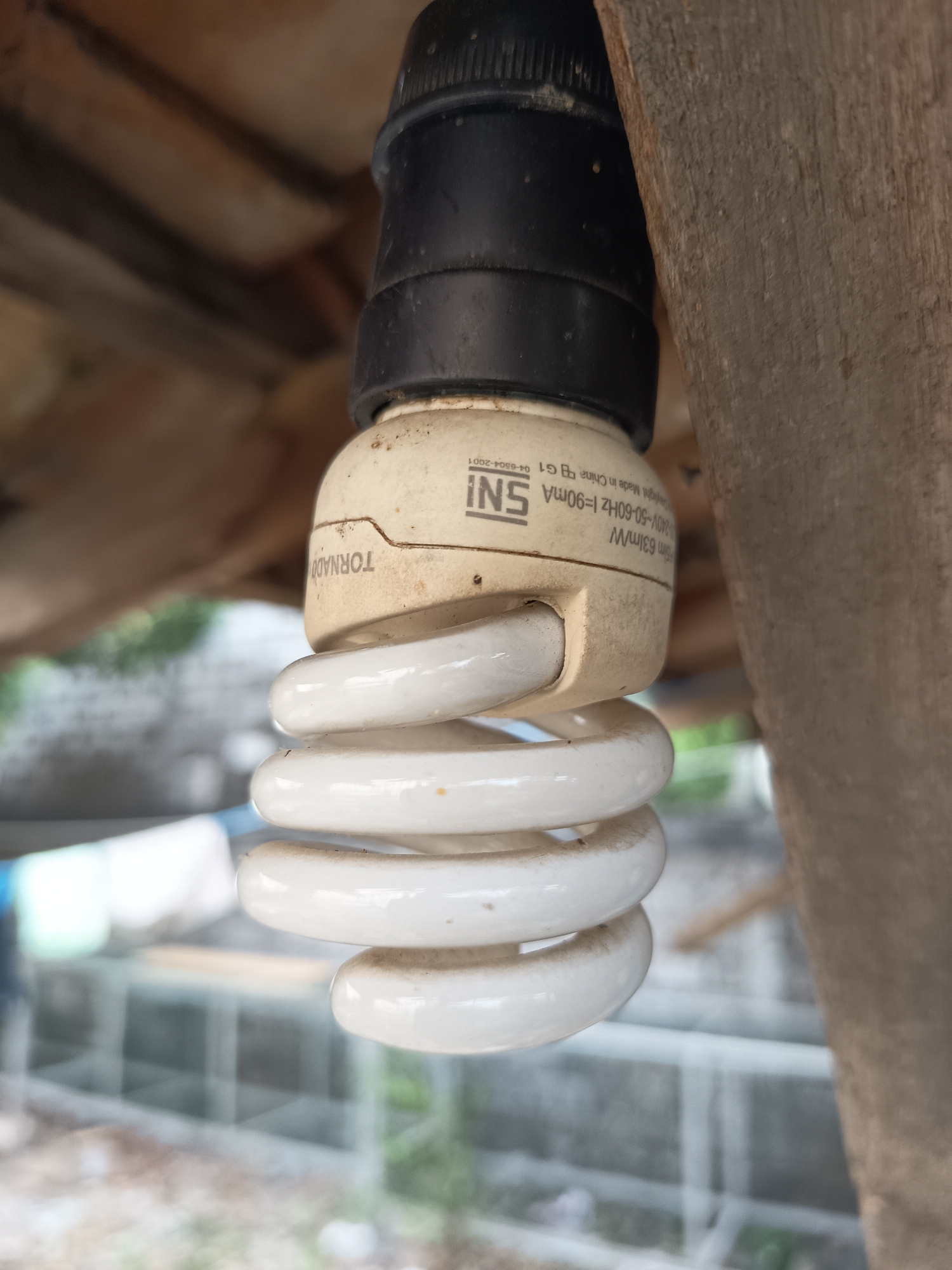 A close-up, eye-level shot shows a compact fluorescent light bulb hanging from a black socket. The bulb is white and spiral-shaped, with a cream-colored base. Text on the base reads "TORNADO," "SNI," and other specifications. The bulb is attached to a wooden beam, with blurred wooden planks above and a blurry outdoor scene in the background, featuring a greenhouse-like structure and laundry hanging. The image was captured on July 5, 2024, at 9:31 AM in Sugio, Indonesia.