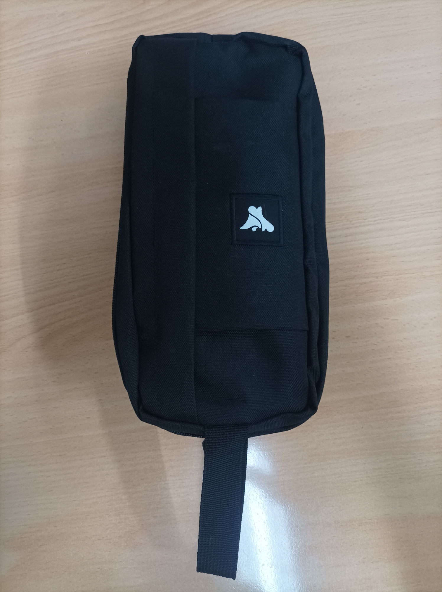 A black fabric pouch with a light blue logo is placed on a light brown wooden surface. The pouch is oriented vertically, with a black strap extending downwards from the bottom center. The logo, located on the upper half of the pouch, appears to be a stylized silhouette of an animal. The wooden surface has a visible wood grain pattern and some reflection from overhead lighting. There are no people or other objects present in the immediate vicinity. The lighting is bright and appears to be coming from above, casting subtle shadows. The image is taken from a top-down perspective.