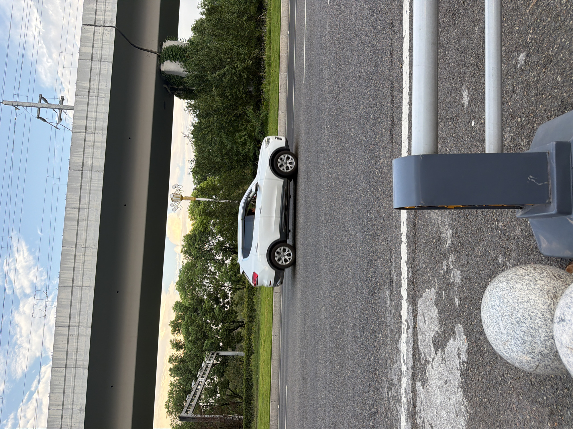 A white SUV drives on a paved road under a concrete overpass. The sky above is blue with scattered clouds, and a wire transmission line stretches across the frame. Trees line the side of the road behind a grassy median. In the foreground, a grey metal barrier with two horizontal bars is visible, along with two large, round stones. The scene appears to be captured during daylight. This location is in the city of Jiangjin, China.