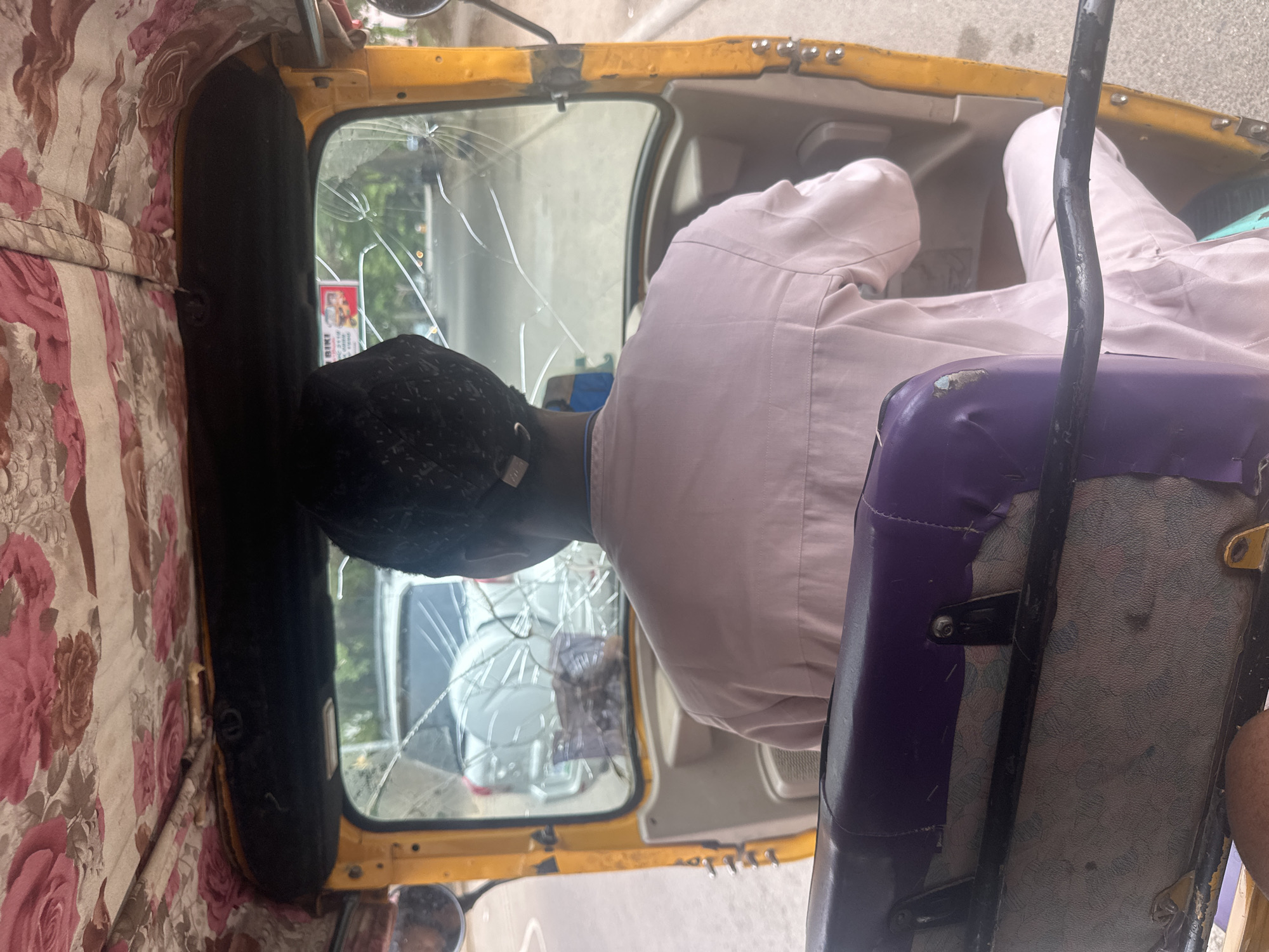 A medium indoor shot shows a person seated inside an auto rickshaw, commonly known as a "Keke" or "Tuk-tuk" in Abuja, Nigeria. The windscreen is cracked, indicating possible damage. The auto rickshaw's interior includes floral patterned wall coverings. The person is wearing a light-colored shirt and a black cap. Seating is visible with a purple and grey color scheme. Exterior view through the damaged windscreen reveals other vehicles and urban elements.