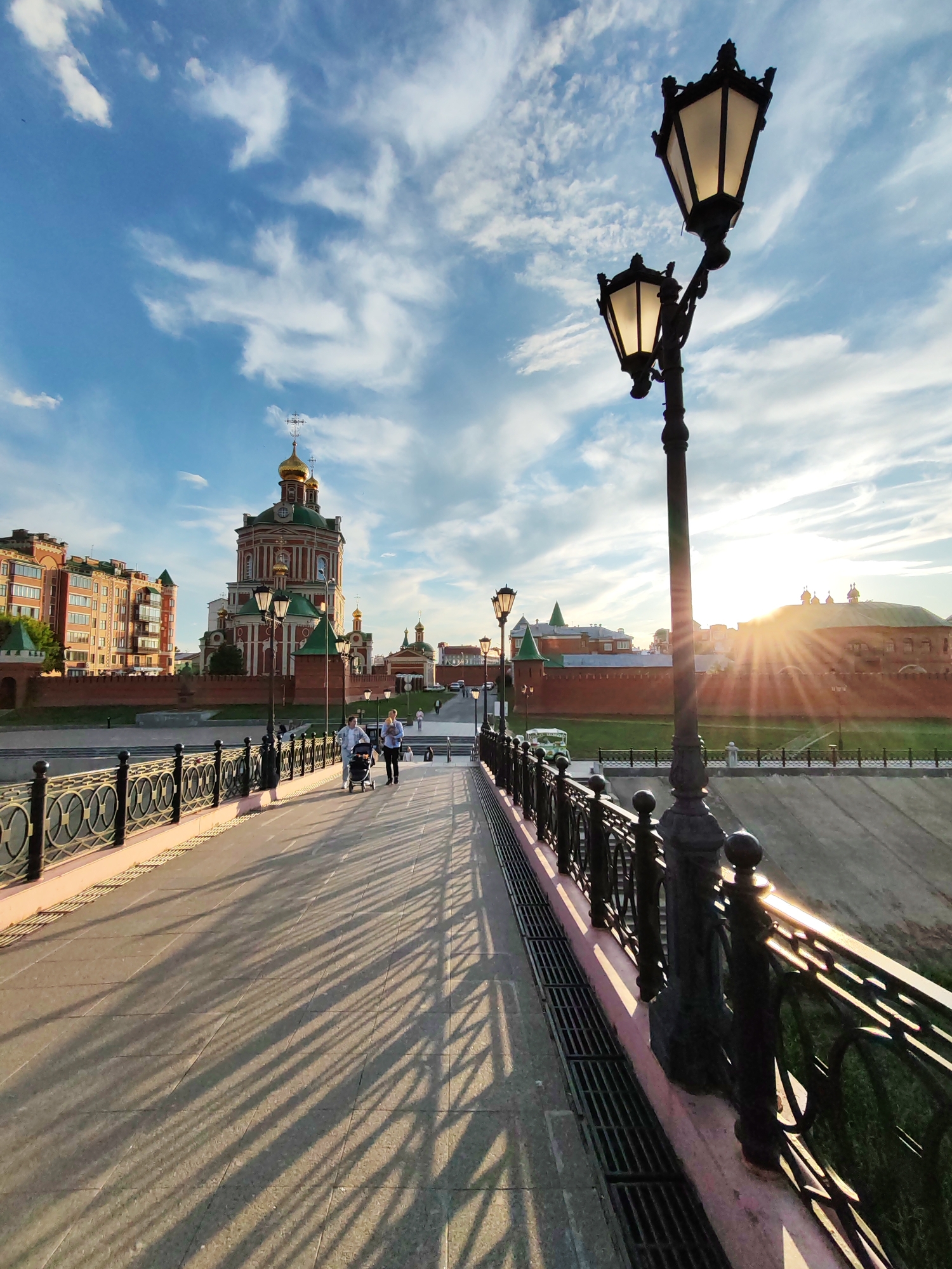 This is a wide-angle, low-angle, eye-level shot of a pedestrian bridge in Yoshkar-Ola, Russia, at sunset. The sun is setting on the right side of the frame, casting long shadows from the lampposts and the bridge railing onto the paved walkway. Two street lamps are visible in the foreground, their warm light illuminating the sky.
In the background, a large, ornate red and green building with golden domes, likely a cathedral or church, is visible, characteristic of Yoshkar-Ola's unique architectural style. To the left of this structure are several modern brick buildings. A red brick wall runs along the embankment, separating the buildings from the river. A few people are walking on the bridge, one pushing a stroller. The sky is a vibrant blue with scattered white clouds, and the sun's rays are breaking through the clouds, creating a dramatic effect. The overall scene conveys a sense of calm and beauty, with the warm light of sunset highlighting the architectural details of the city.